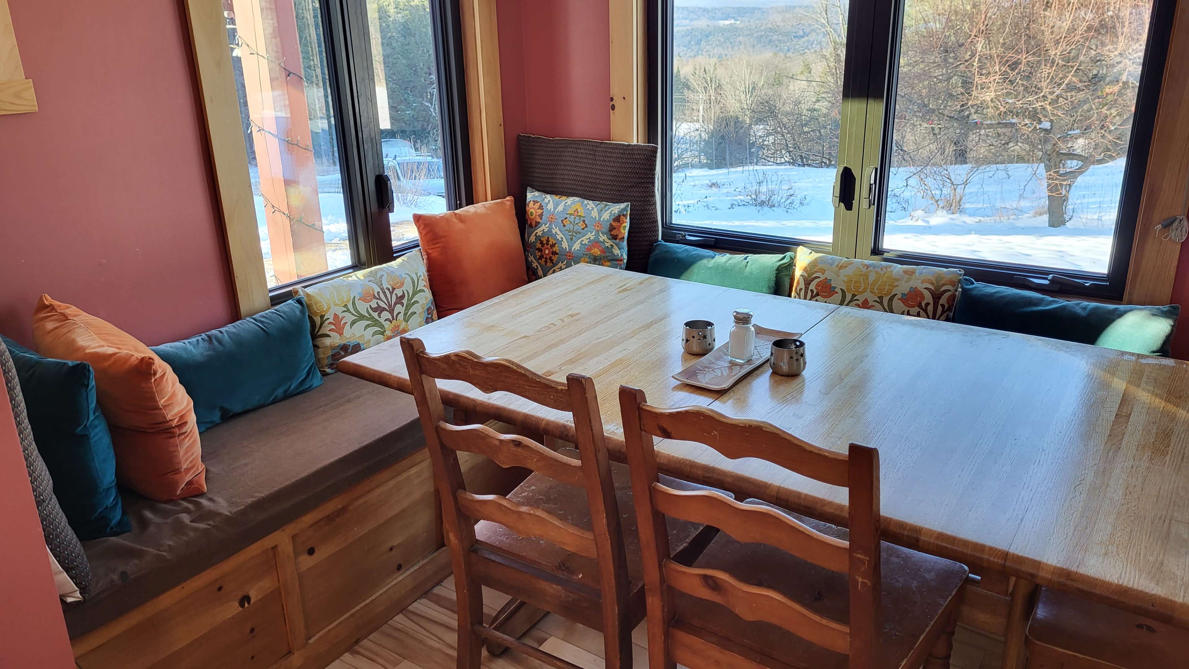 A wooden dining table with several chairs is positioned near large windows, surrounded by colorful cushions, overlooking a snowy landscape.