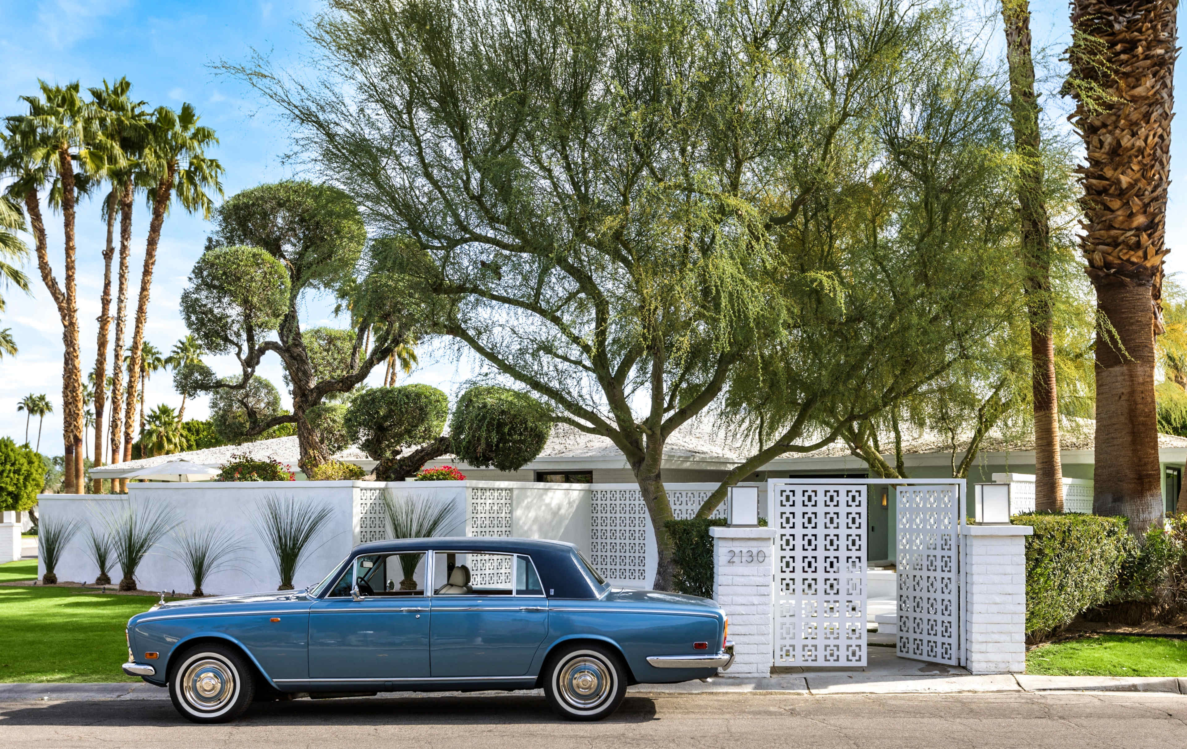 A vintage blue car is parked in front of a well-maintained house surrounded by palm trees and a decorative white fence.