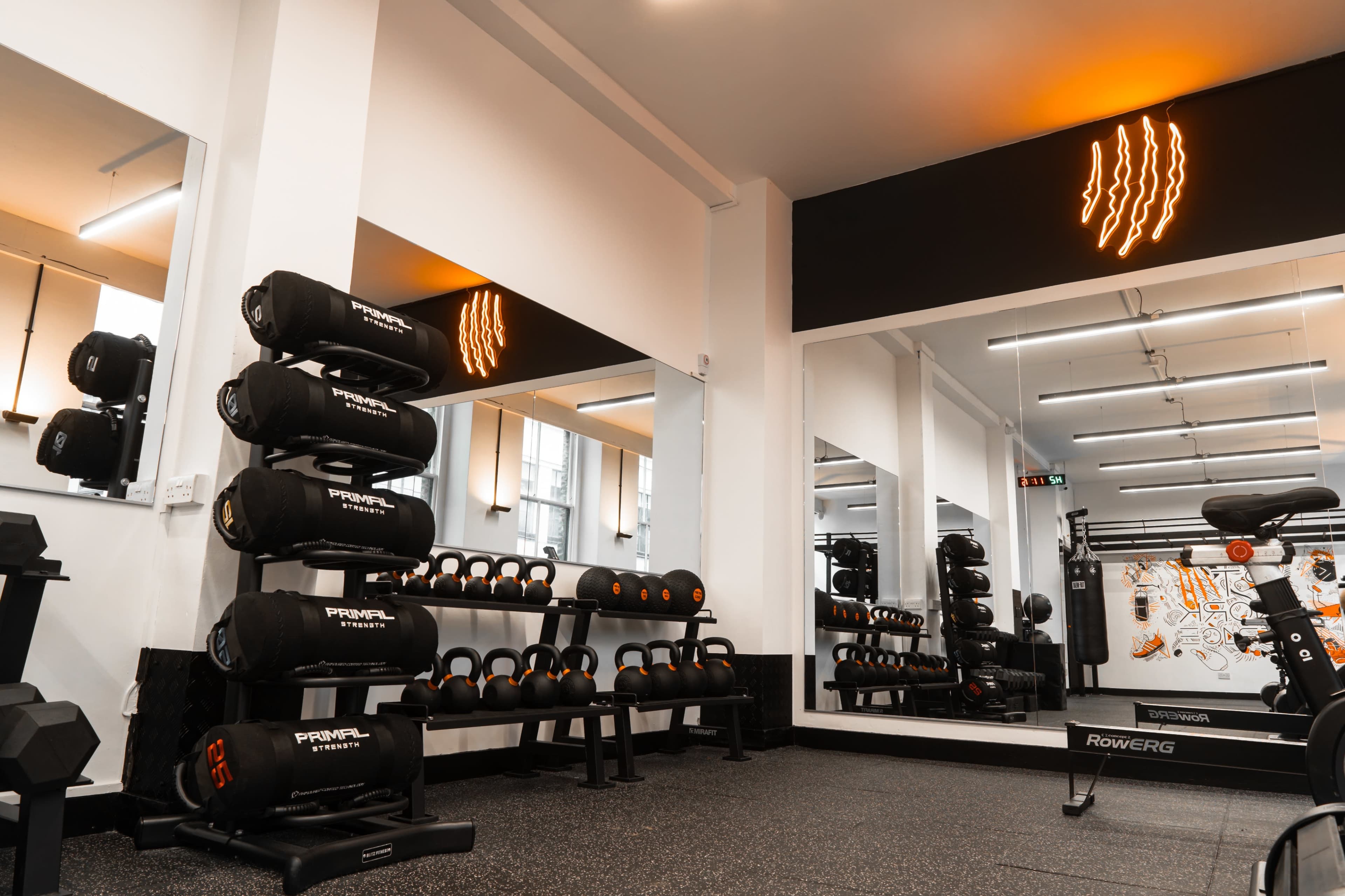 The image shows a modern gym interior featuring a stacked rack of black dumbbells, a weight bench, and a rowing machine with a large mirror and neon wall art in the background.