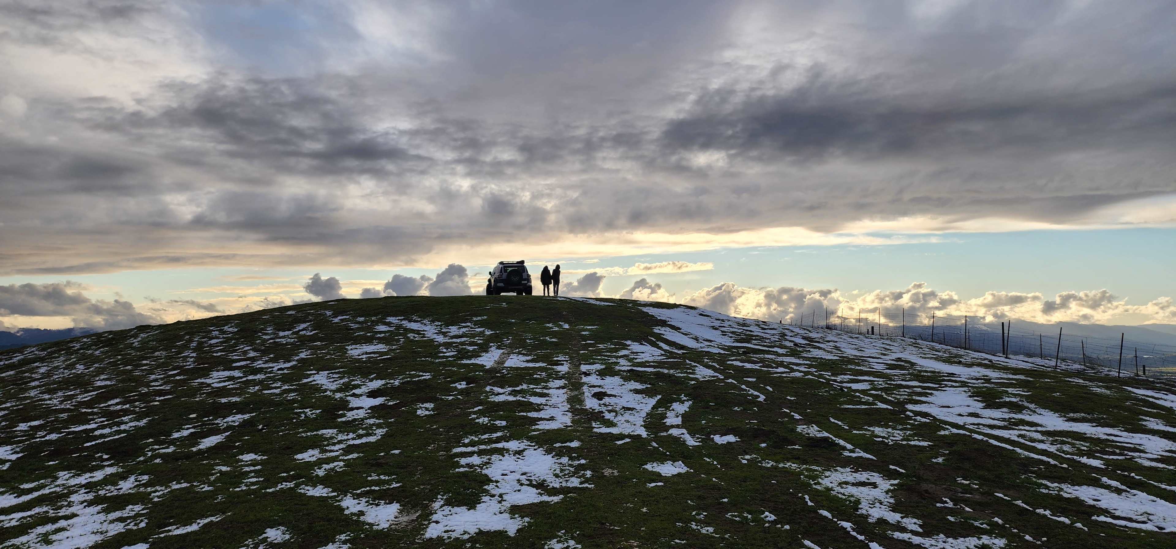 A vehicle and two people stand on a snow-covered hill under a cloudy sky.