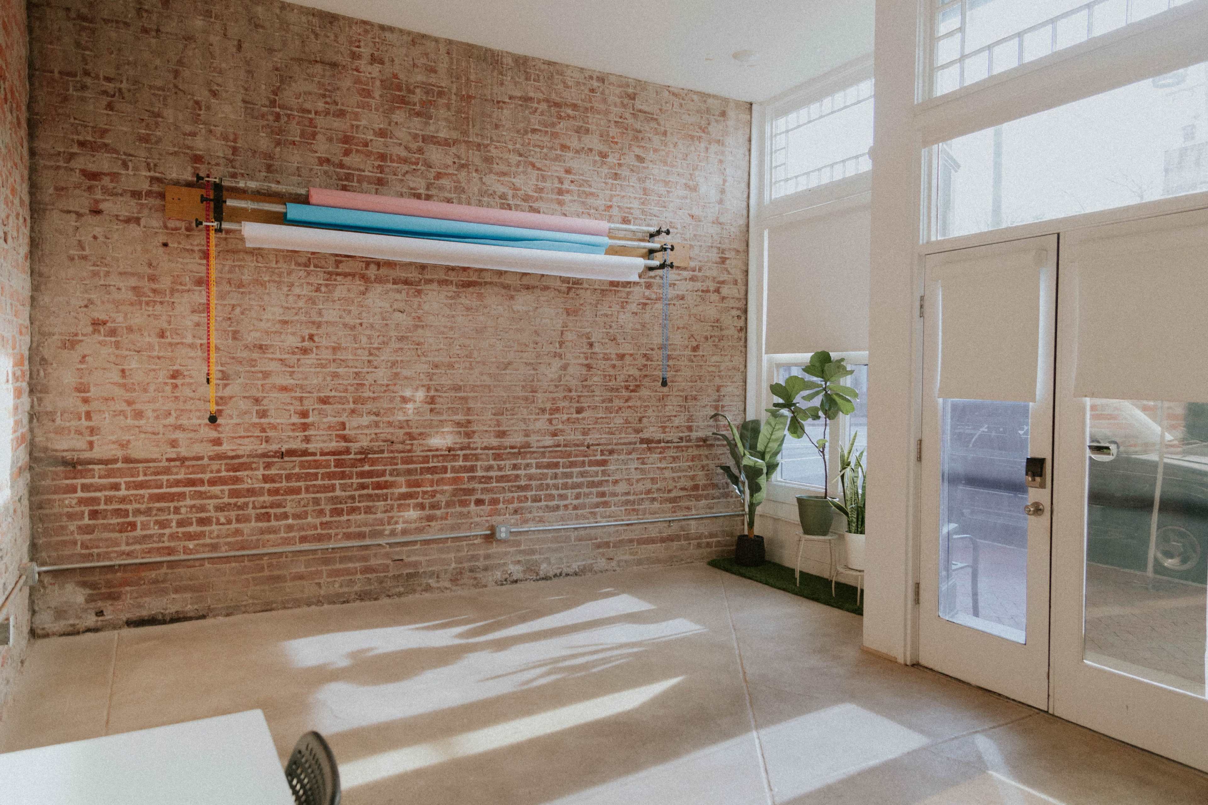 The image shows a bright interior of a room with exposed brick walls, large windows, and a roll of colorful paper mounted on the wall, alongside a potted plant and a door leading outside.