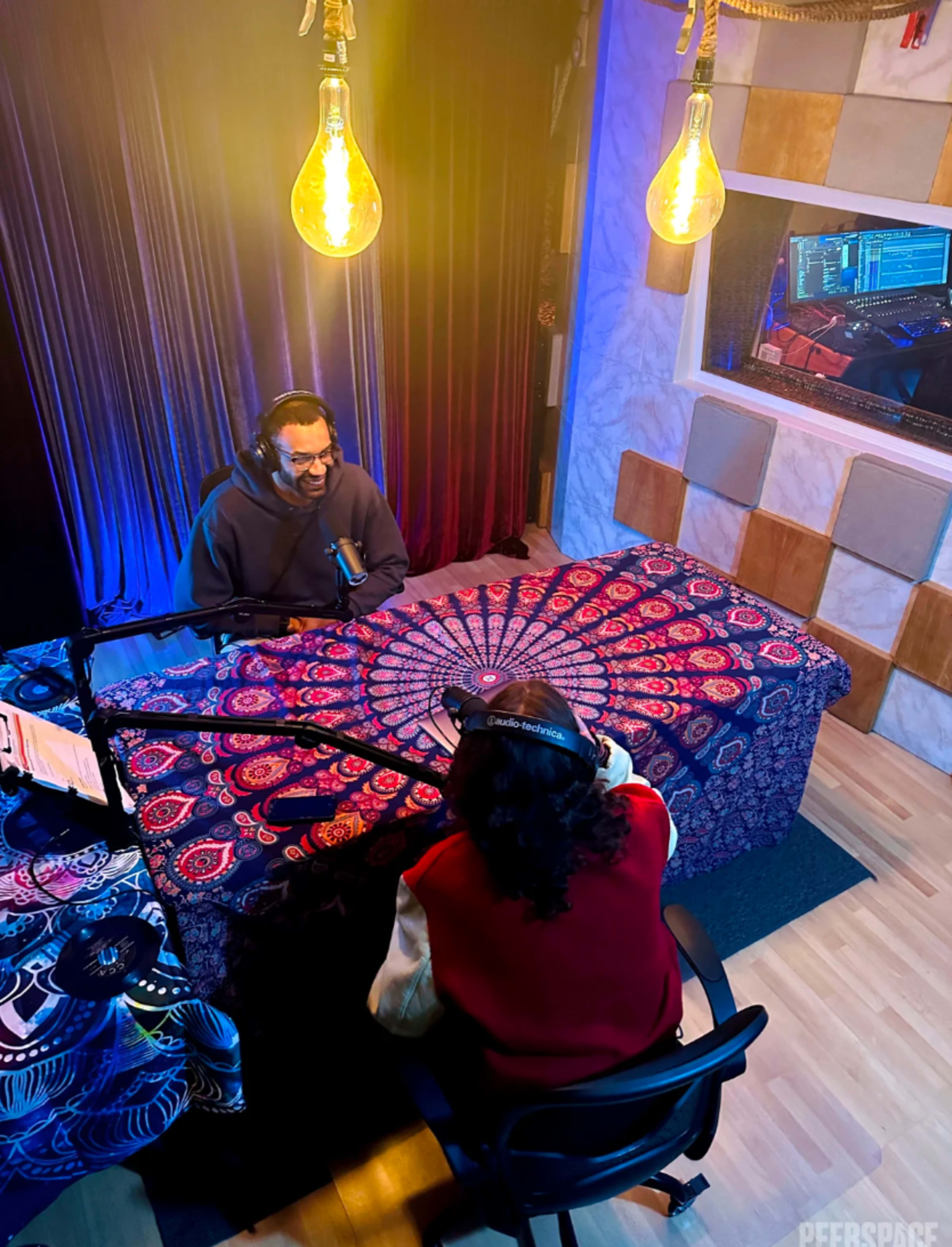 Two people sit at a table covered with a patterned cloth in a recording studio, speaking into microphones with warm light bulbs hanging above.