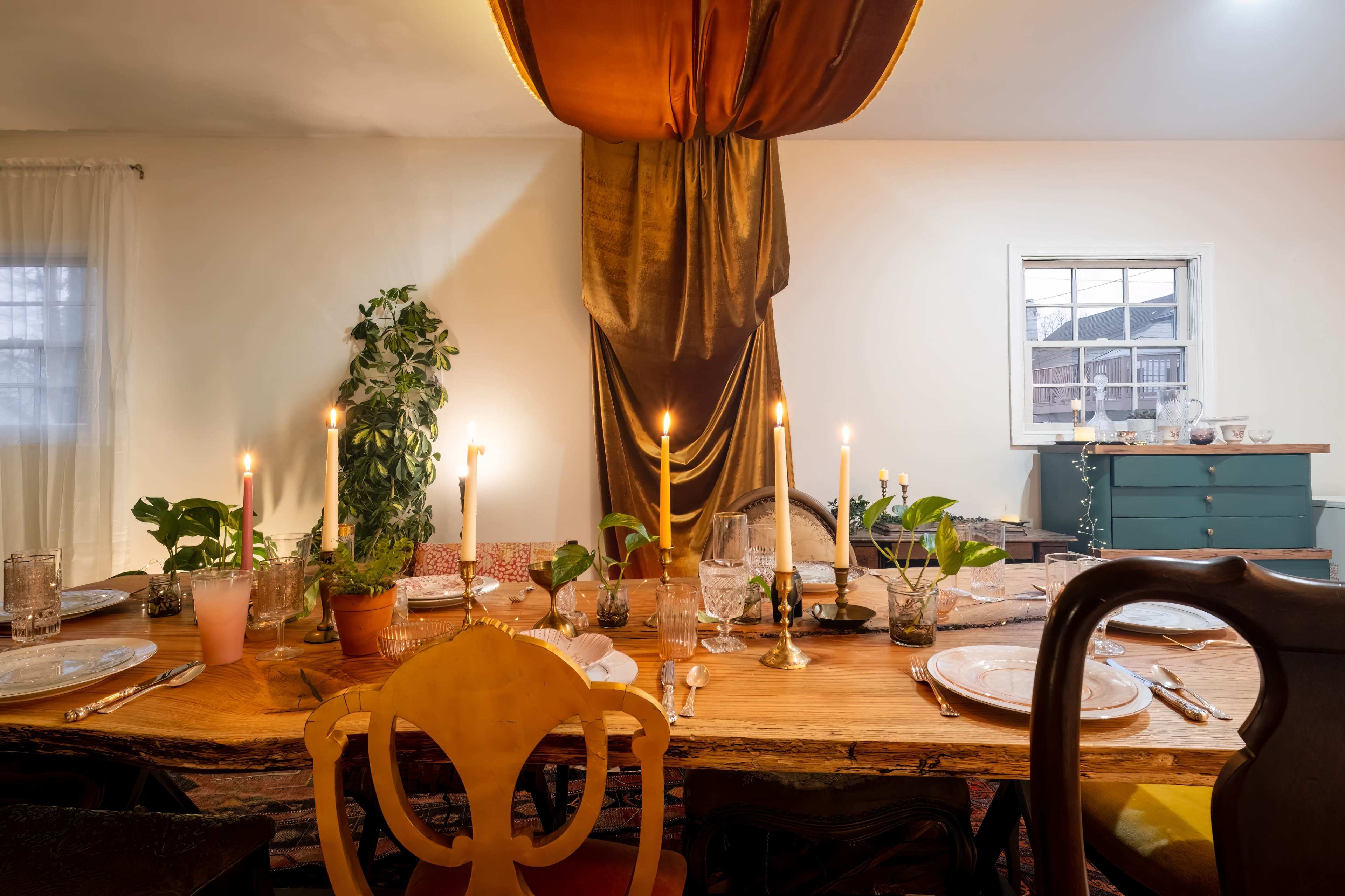 A dining table is set for a meal, adorned with candles, plants, and elegant tableware, beneath a decorative drapery.