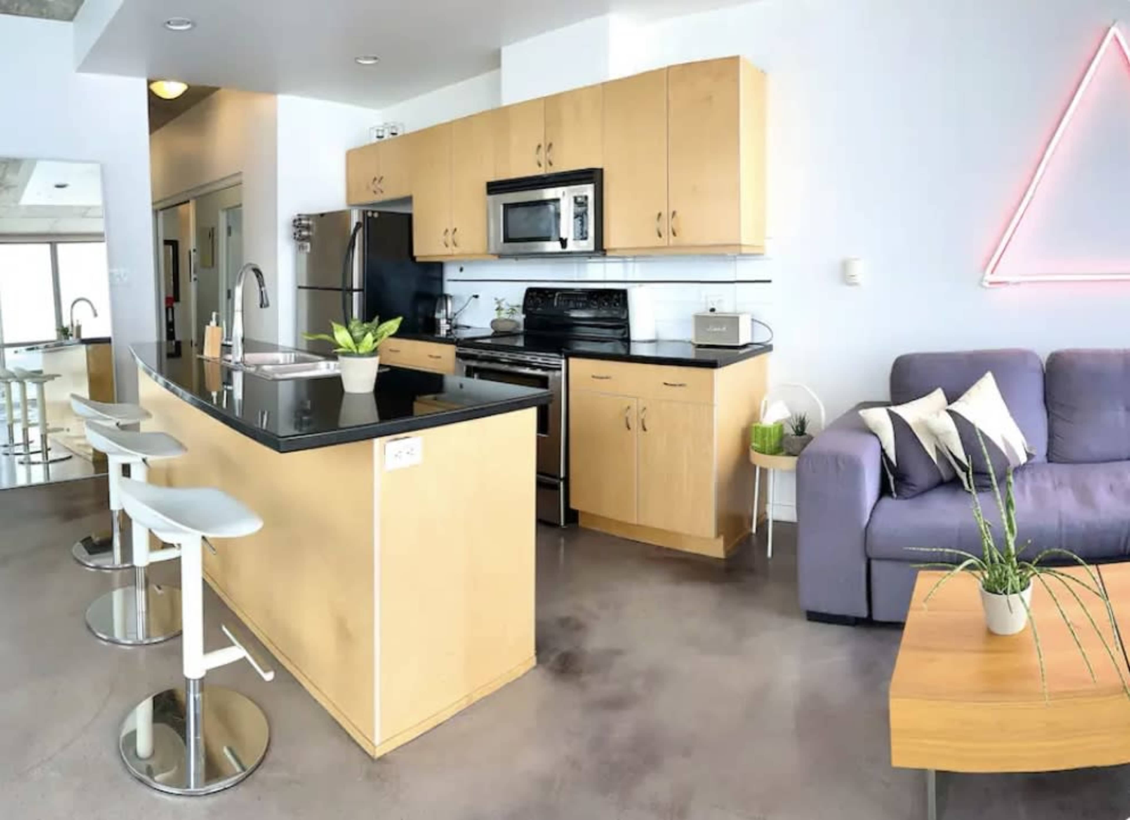 A modern kitchen and living area with light wood cabinetry, stainless steel appliances, and a gray sofa.
