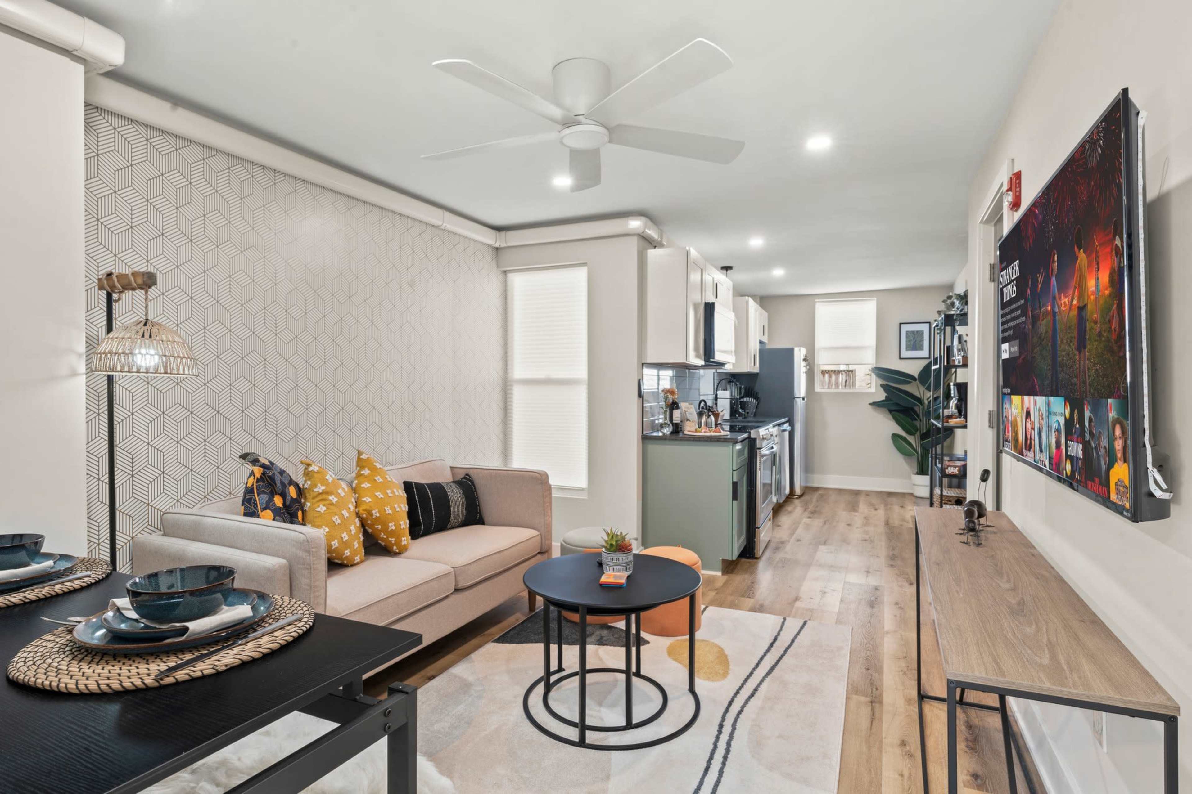 A modern living room with a sofa, coffee table, kitchen area, and a large wall-mounted television.