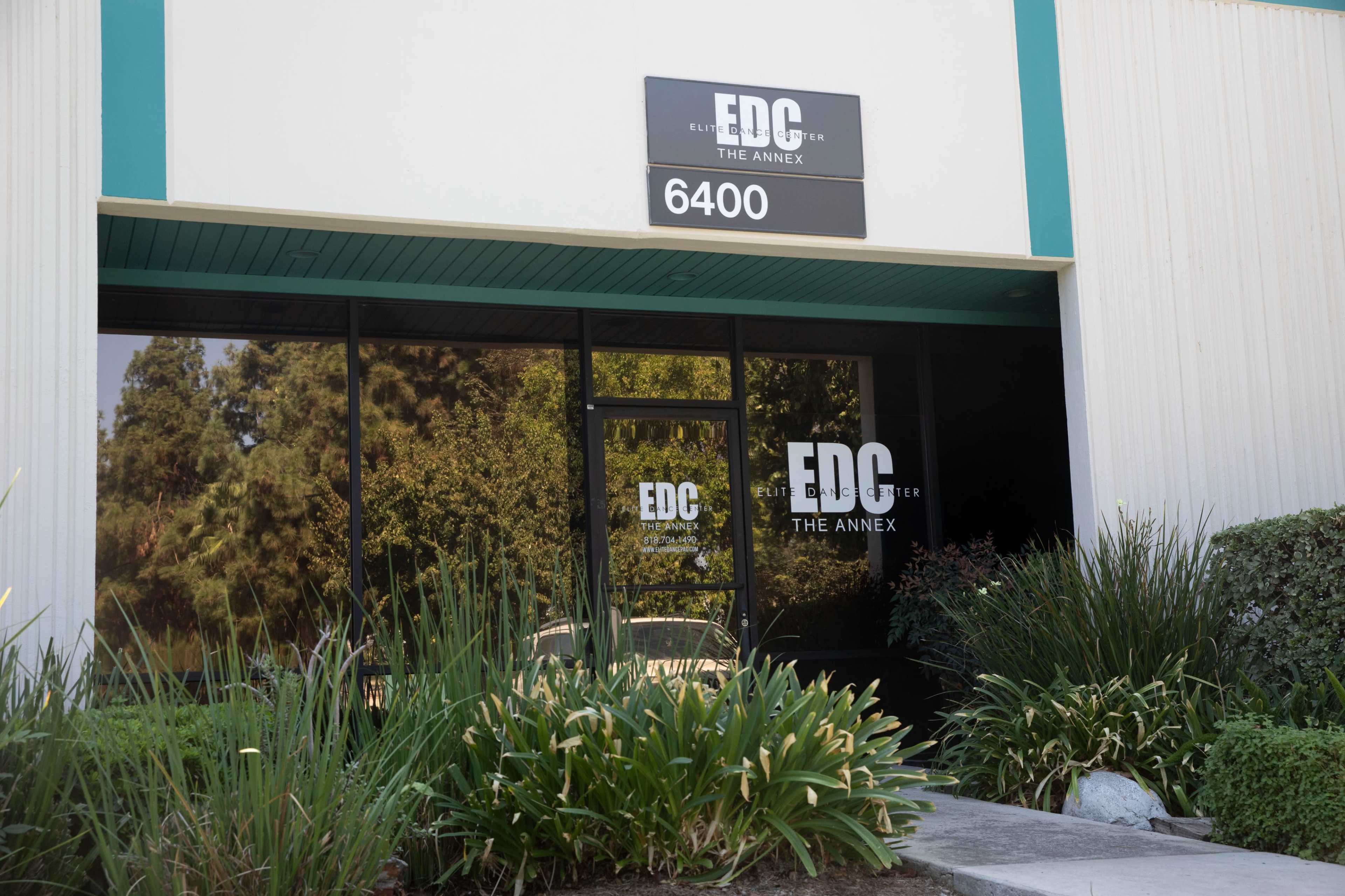 A modern building entrance with a sign reading "EDC THE ANNEX" and a large glass door surrounded by greenery.