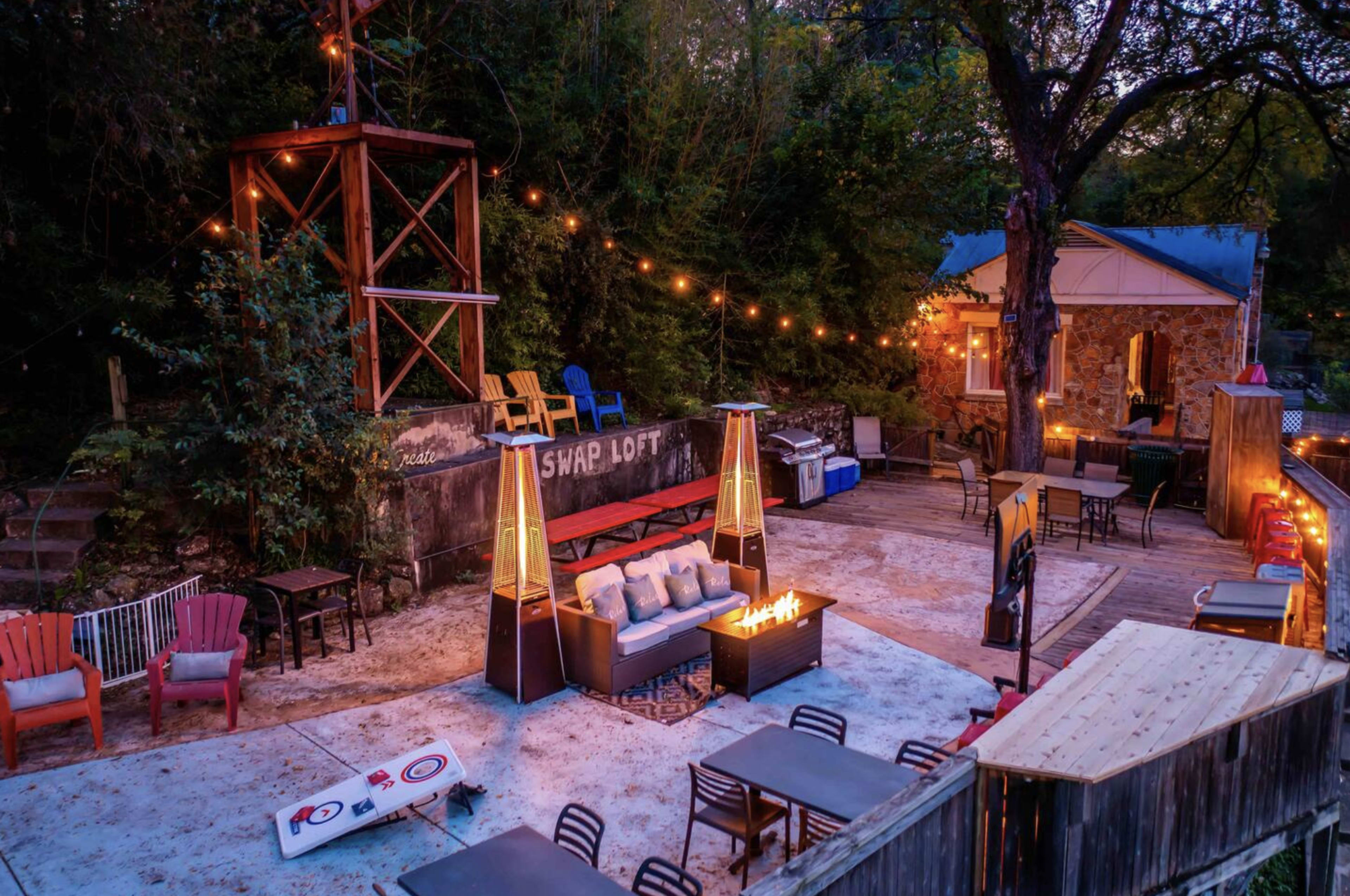 The image depicts an outdoor area with seating, string lights, a fire pit, and a wooden deck surrounded by trees and a building.