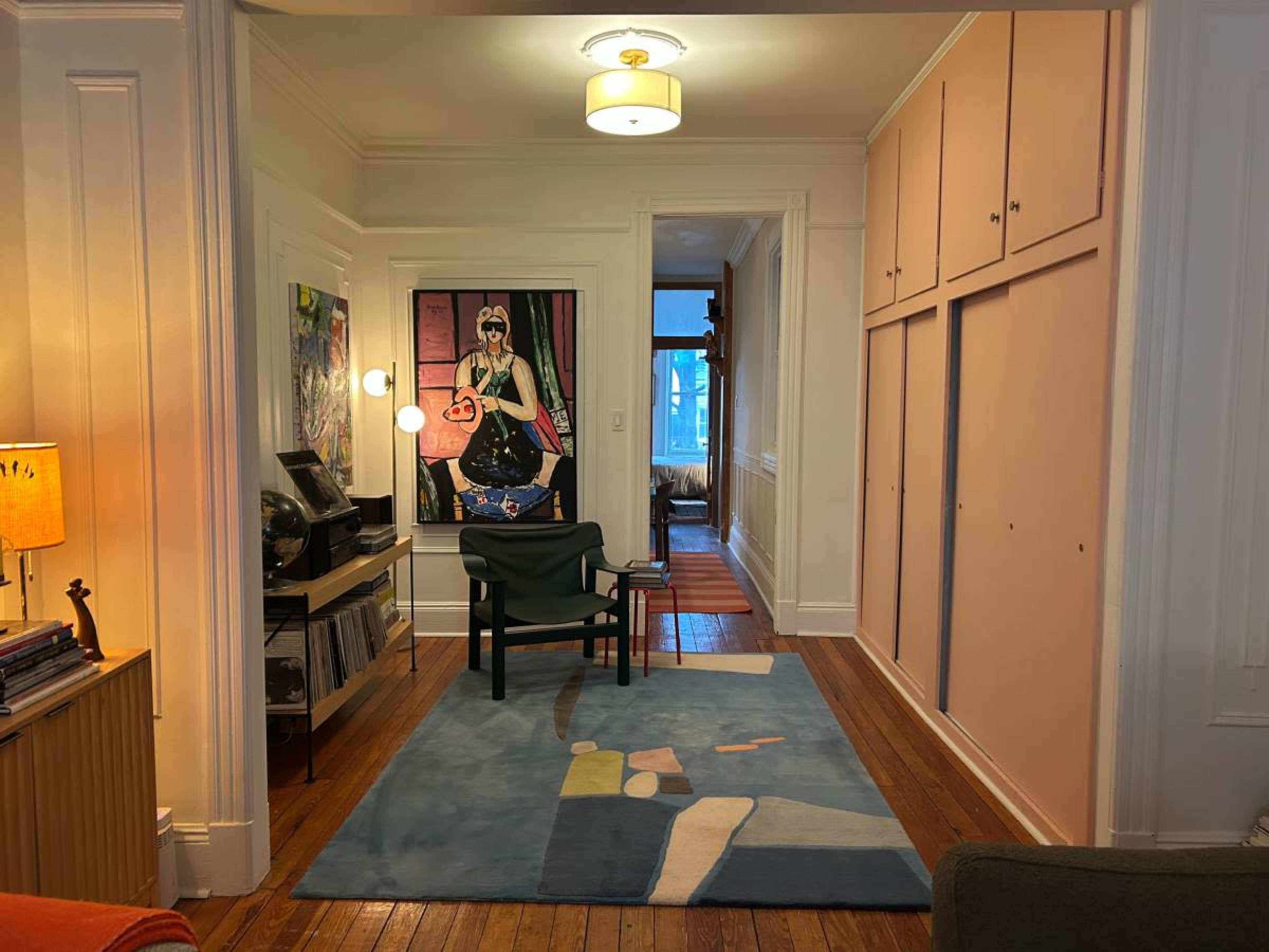 The image shows a hallway with a colorful rug, a chair, and art on the wall, leading to another room with light coming in through a window.