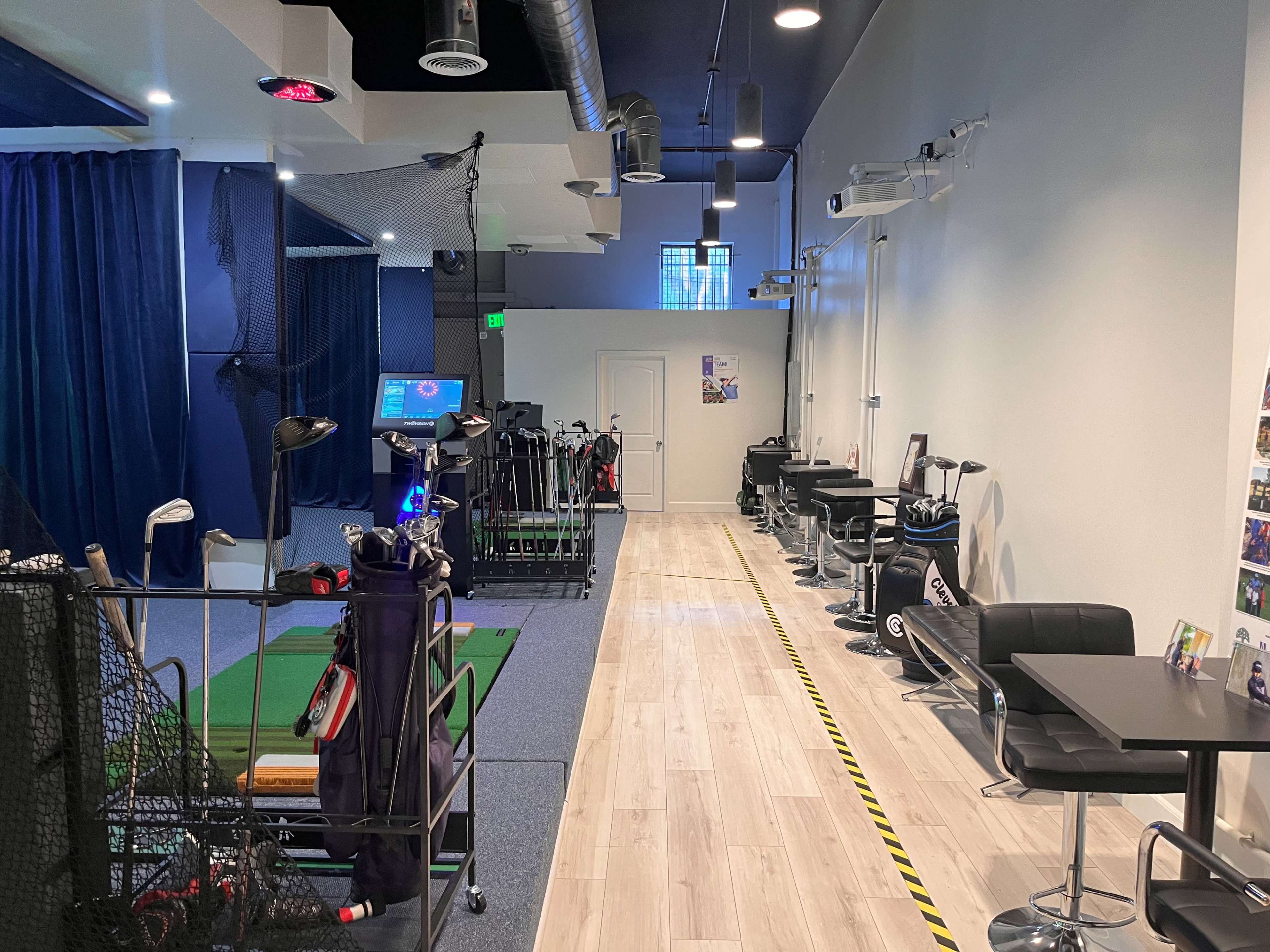 An indoor golf simulation facility with a carpeted area for practice on one side and tables and chairs along the opposite wall.