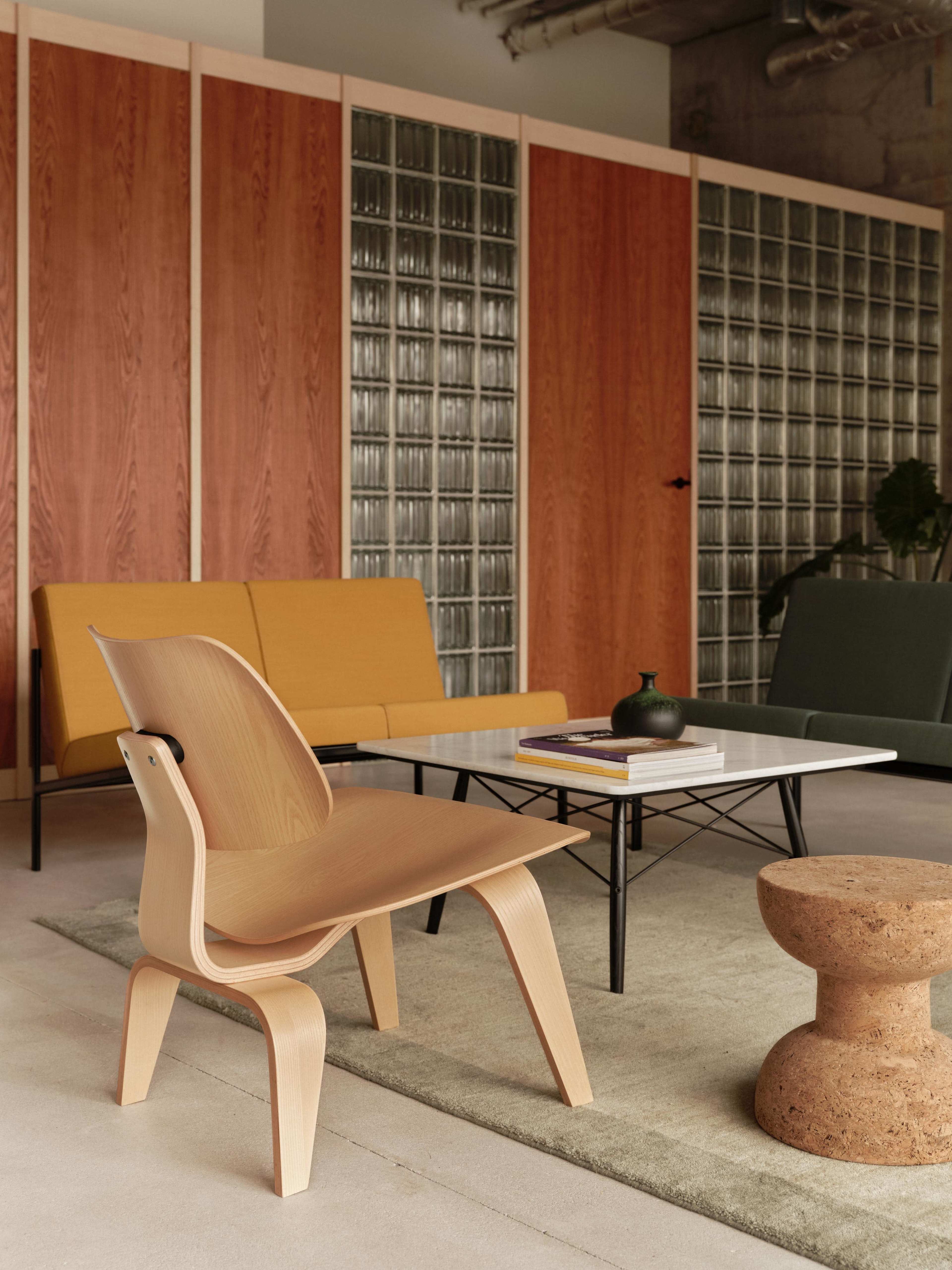 The image shows a modern interior with a wooden chair, a low table with a book and a decorative vase, and two upholstered couches in a spacious room featuring glass block walls.