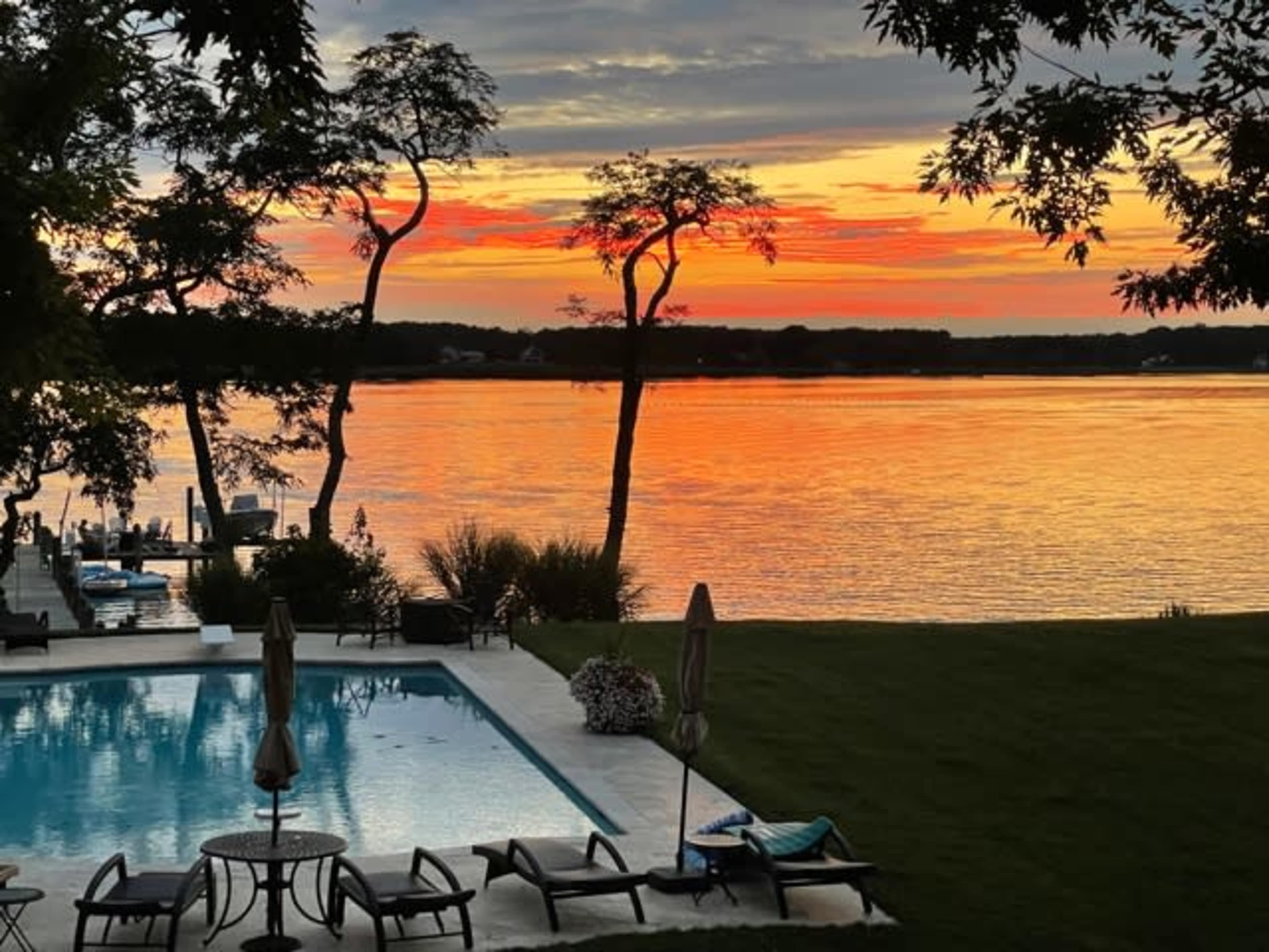A serene view of a lake at sunset, reflecting vibrant colors, with a swimming pool and lounge chairs in the foreground.