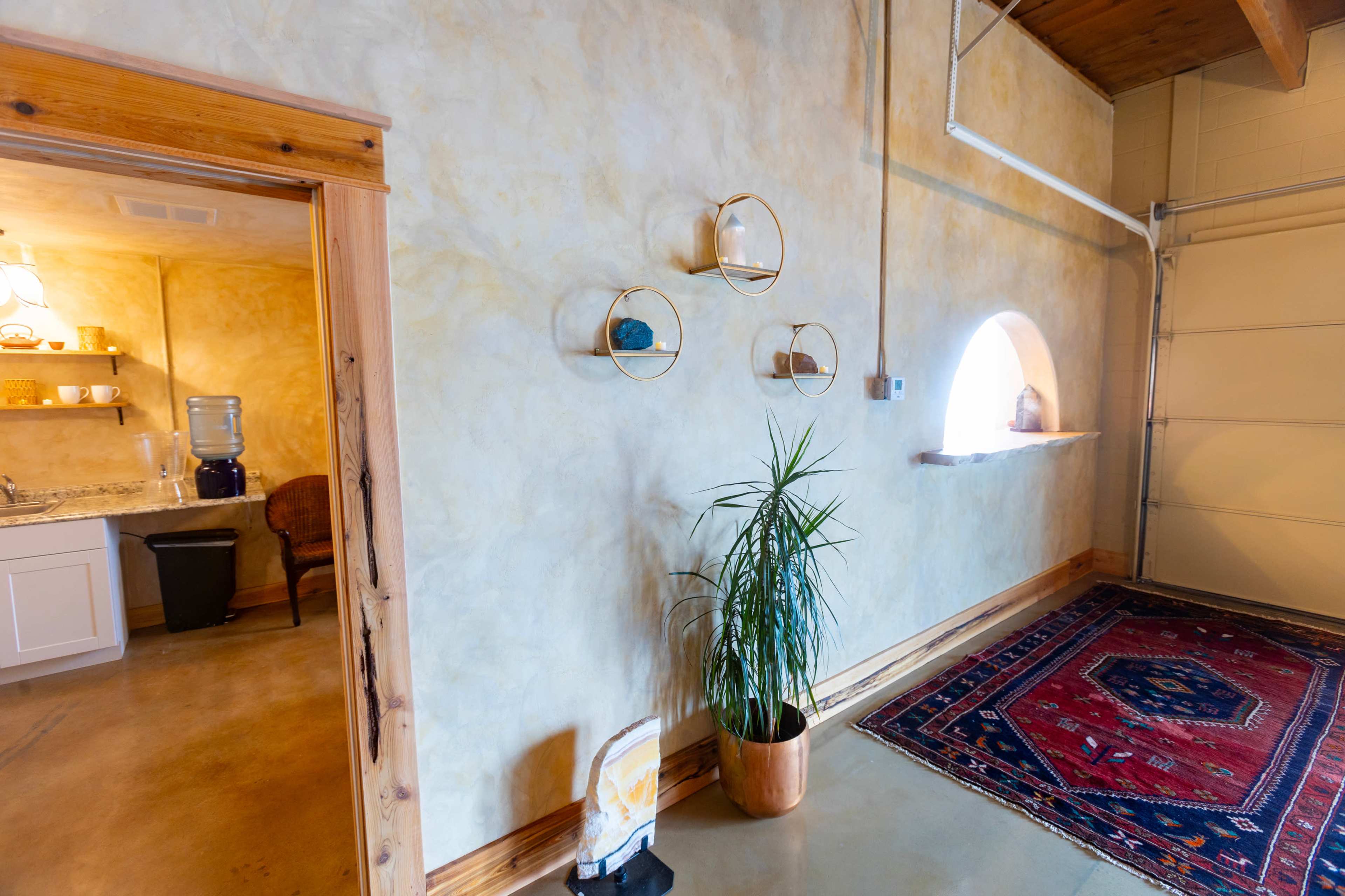 A room with a textured yellow wall, a decorative plant in a pot, three circular wall shelves, a window arch, and a patterned rug on the floor.