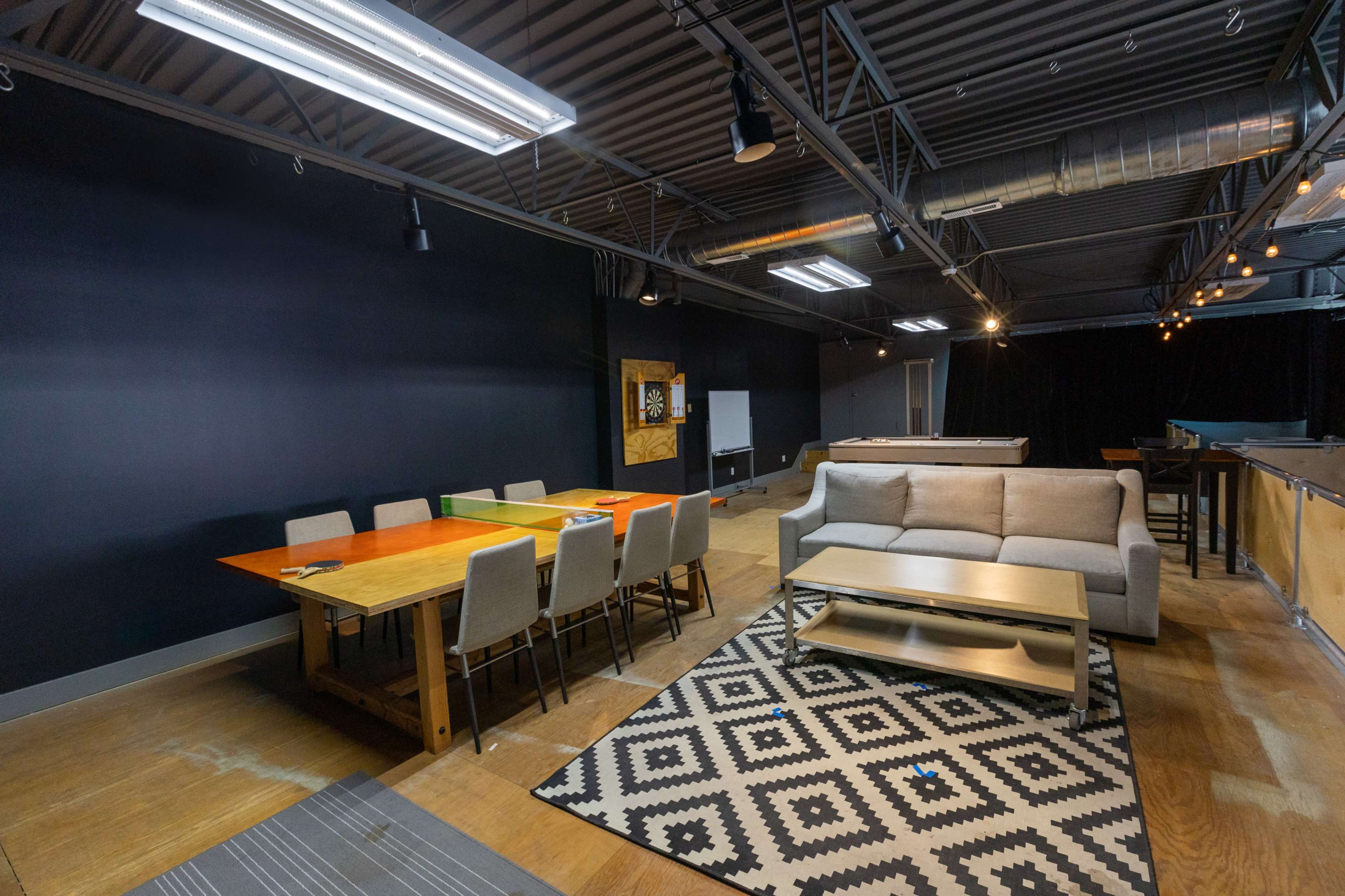 A spacious room with a wooden dining table surrounded by chairs, a sofa, and a patterned rug, all under industrial-style lighting.