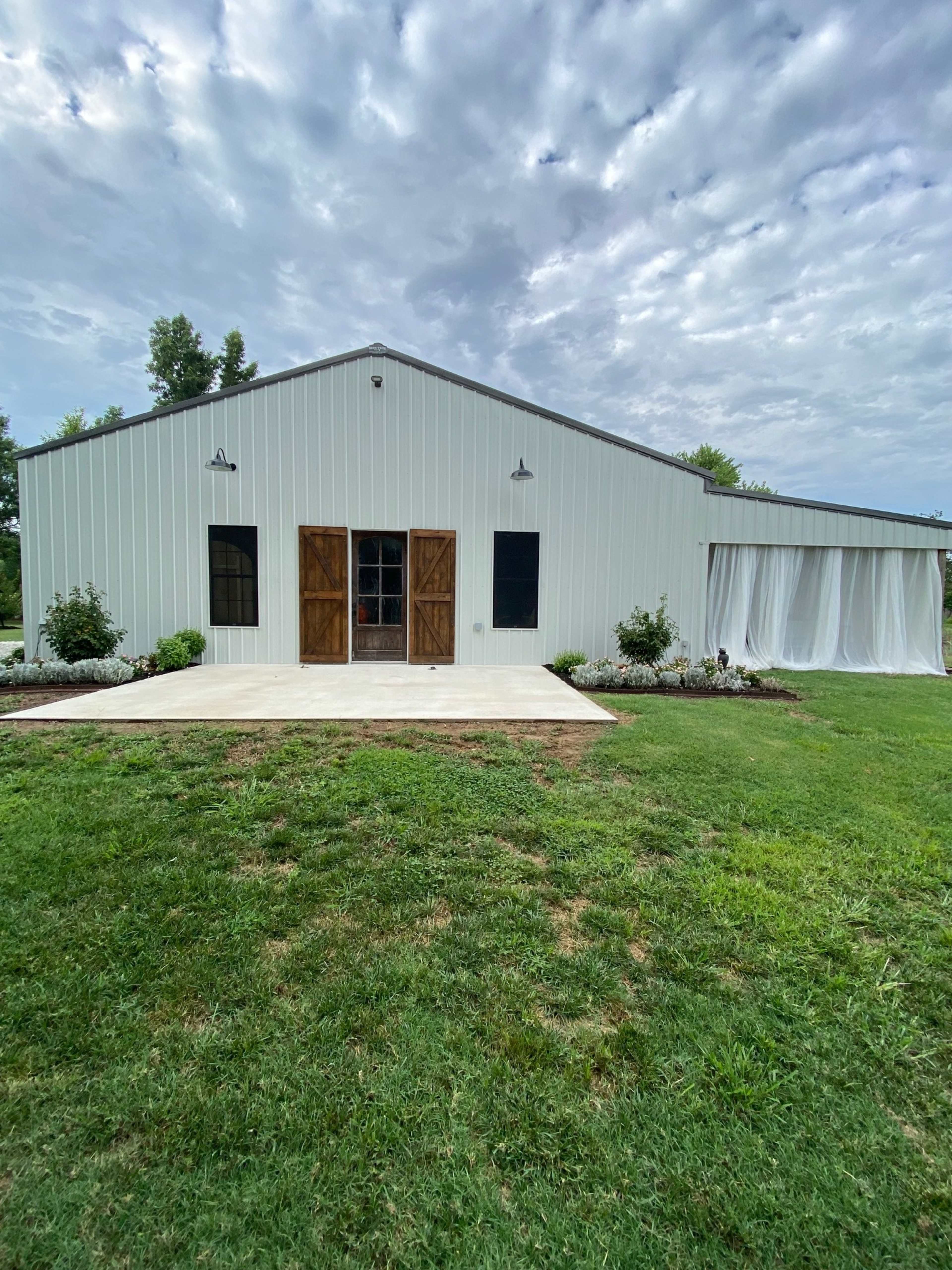 A large white metal building with dual wooden doors and windows stands on a grassy area, featuring a covered patio with landscaping in front.