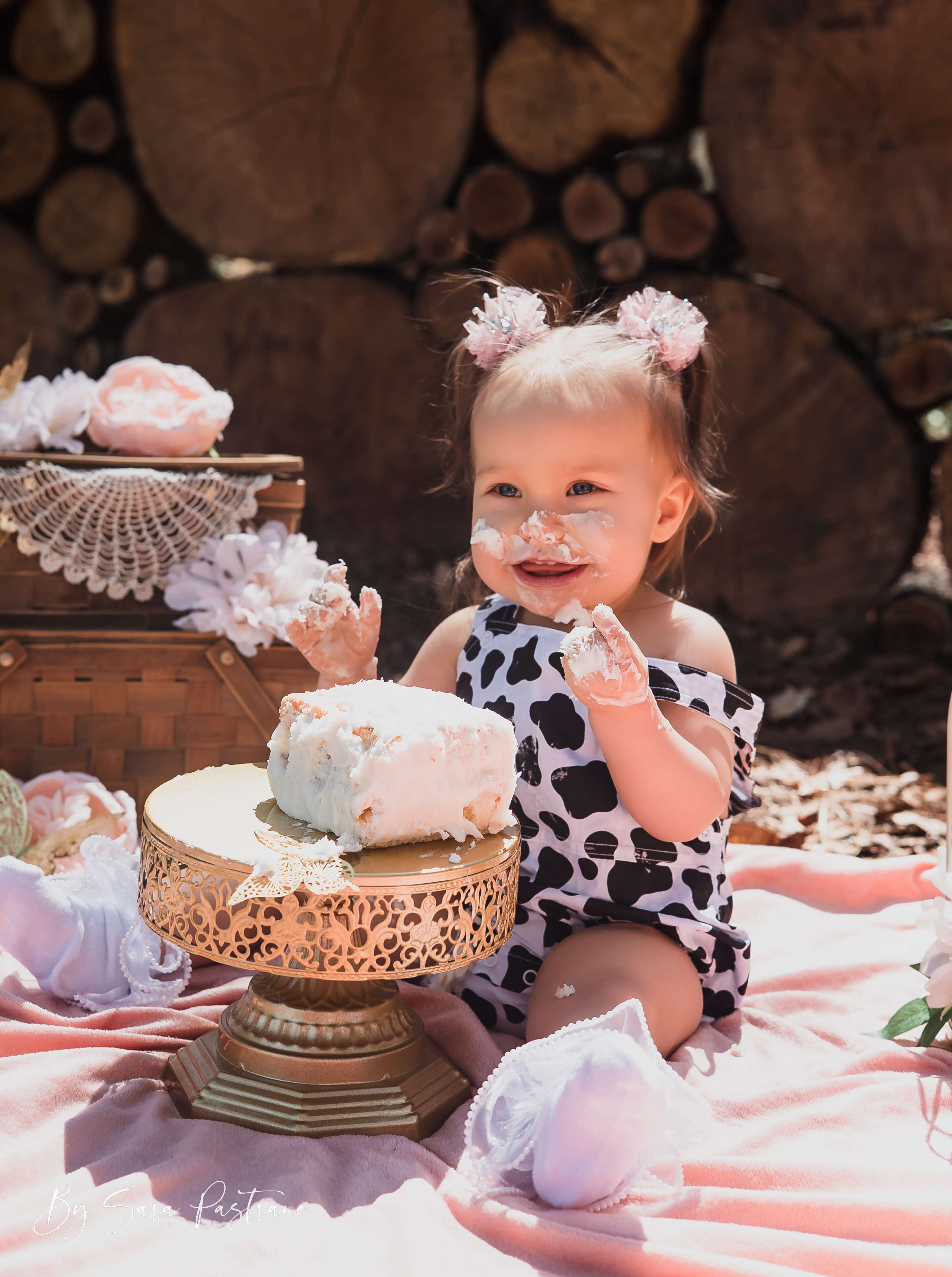 A toddler wearing a black and white cow-print outfit sits on a blanket surrounded by decorative items, enjoying a large slice of cake.