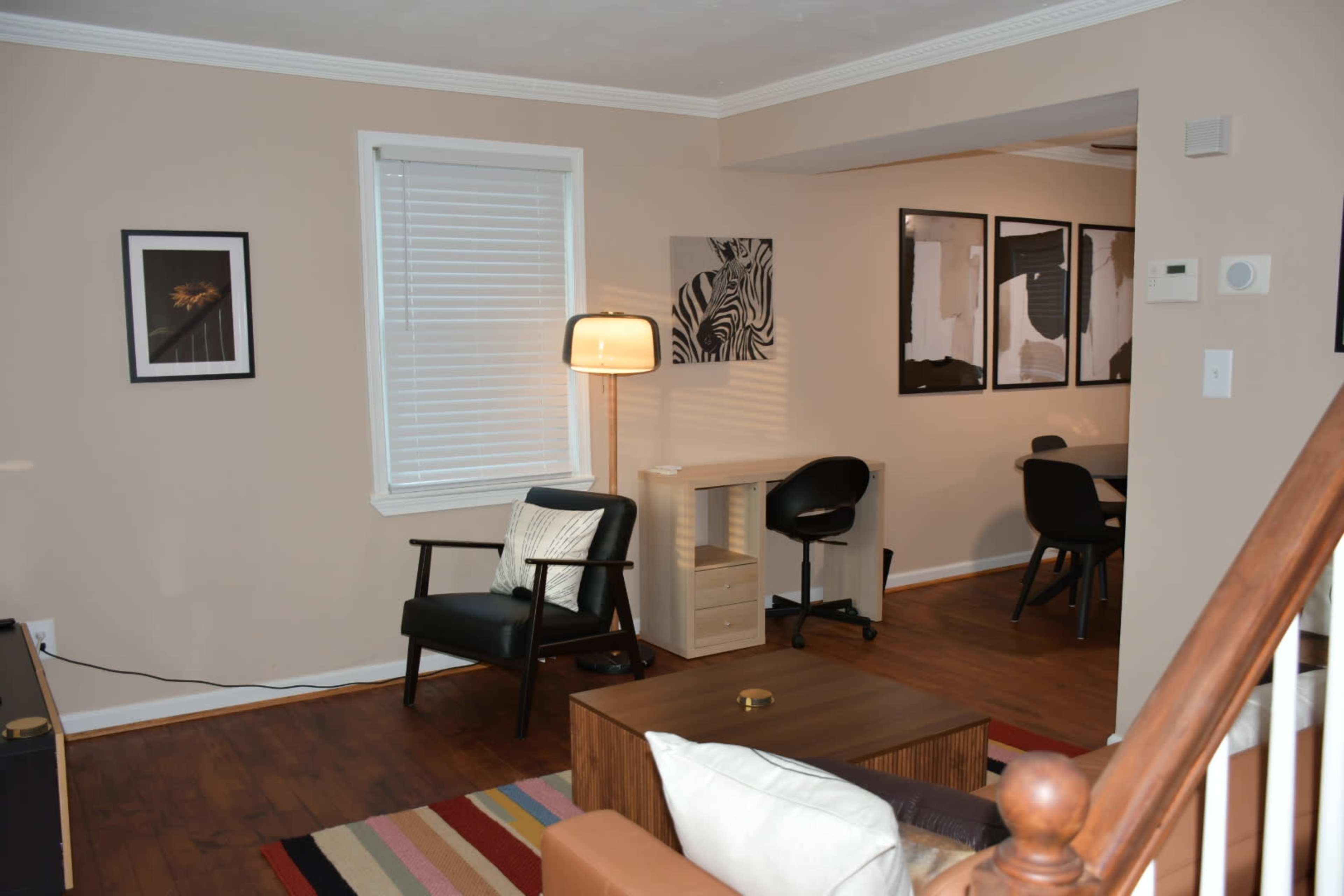 The image shows a modern living room with a lamp, a black chair, a small wooden table, and a dining area featuring black chairs and framed artwork on the walls.