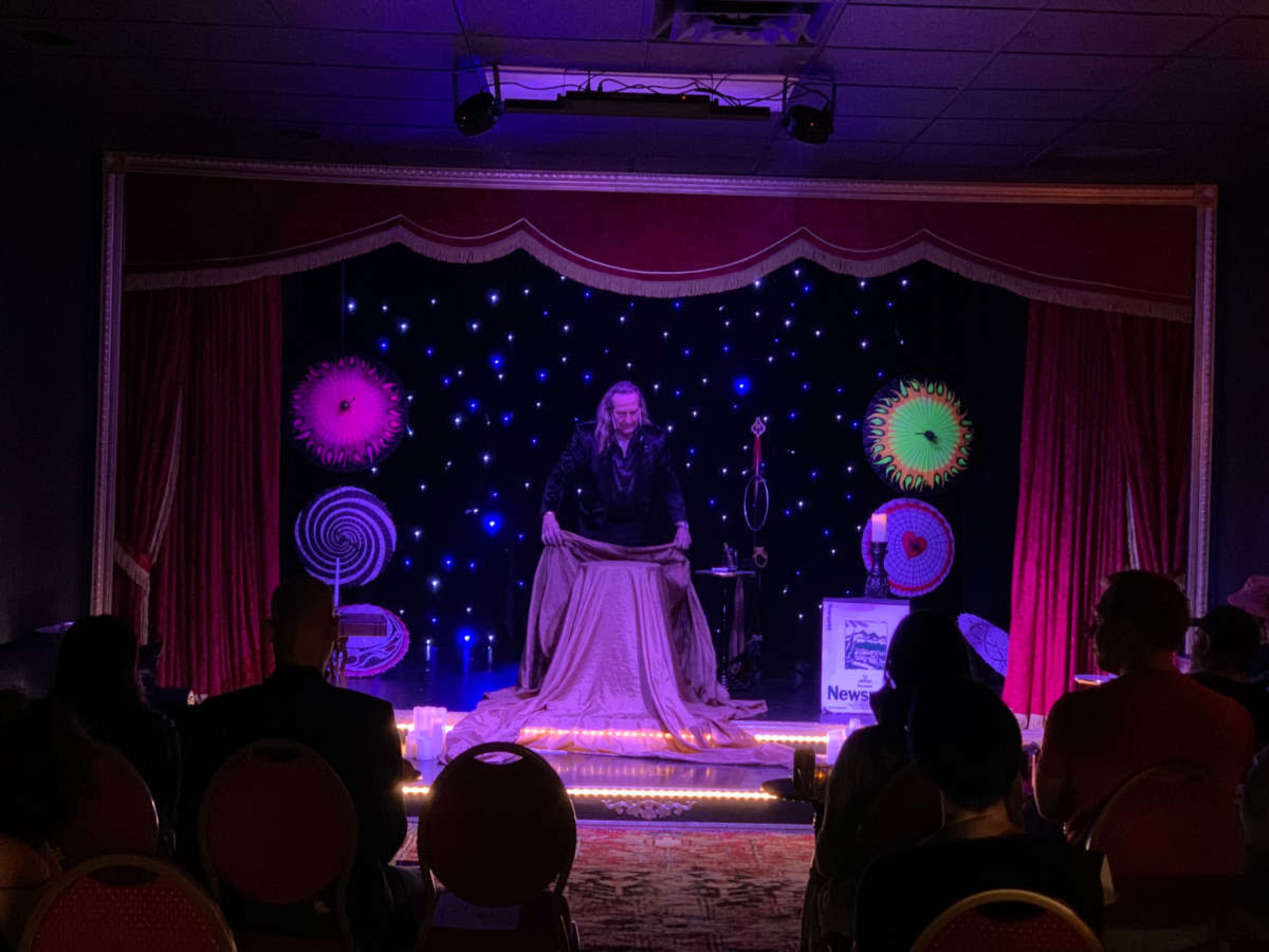 A performer dressed in dark clothing stands on stage, draped in fabric, under colorful stage lights and a backdrop of twinkling stars.