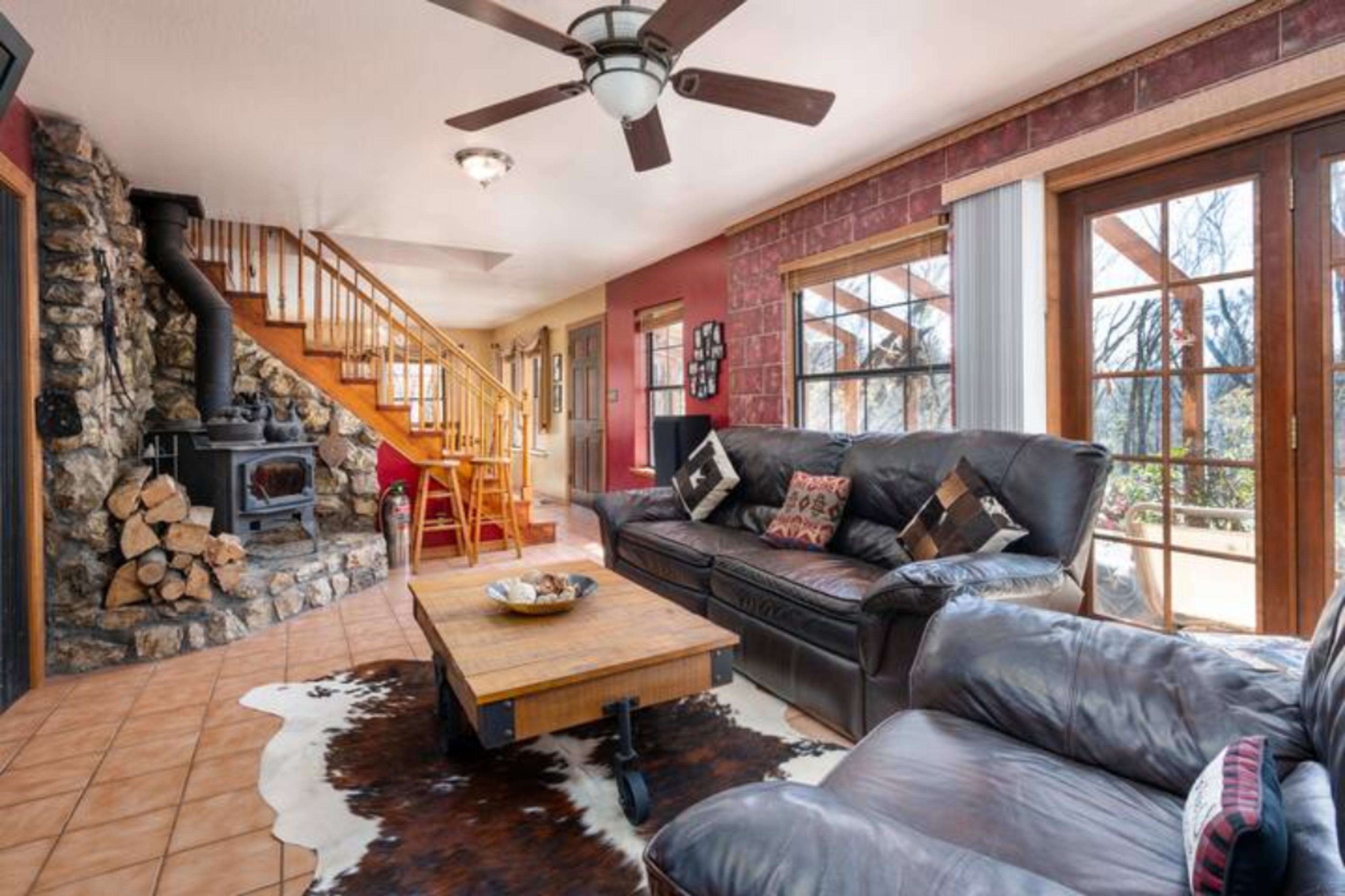 A living room with a stone fireplace, a wooden staircase, a coffee table, and large windows leading to a deck.