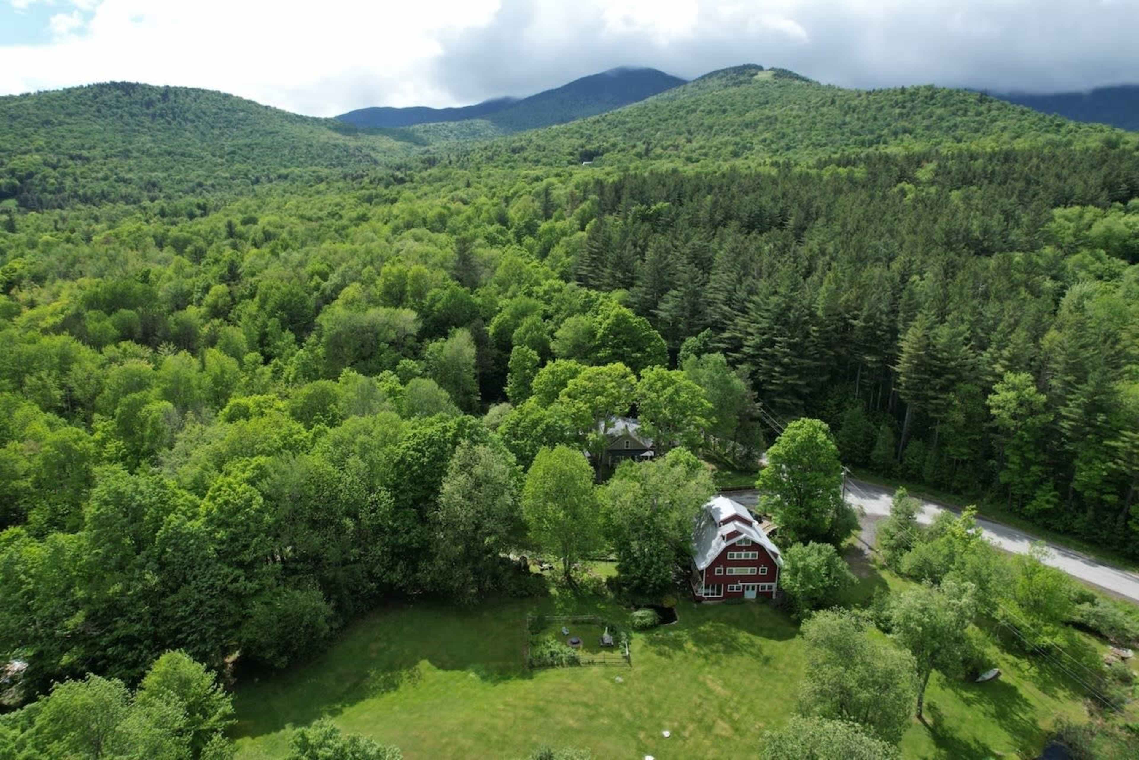 An aerial view shows a red house surrounded by dense greenery and mountains in the background.