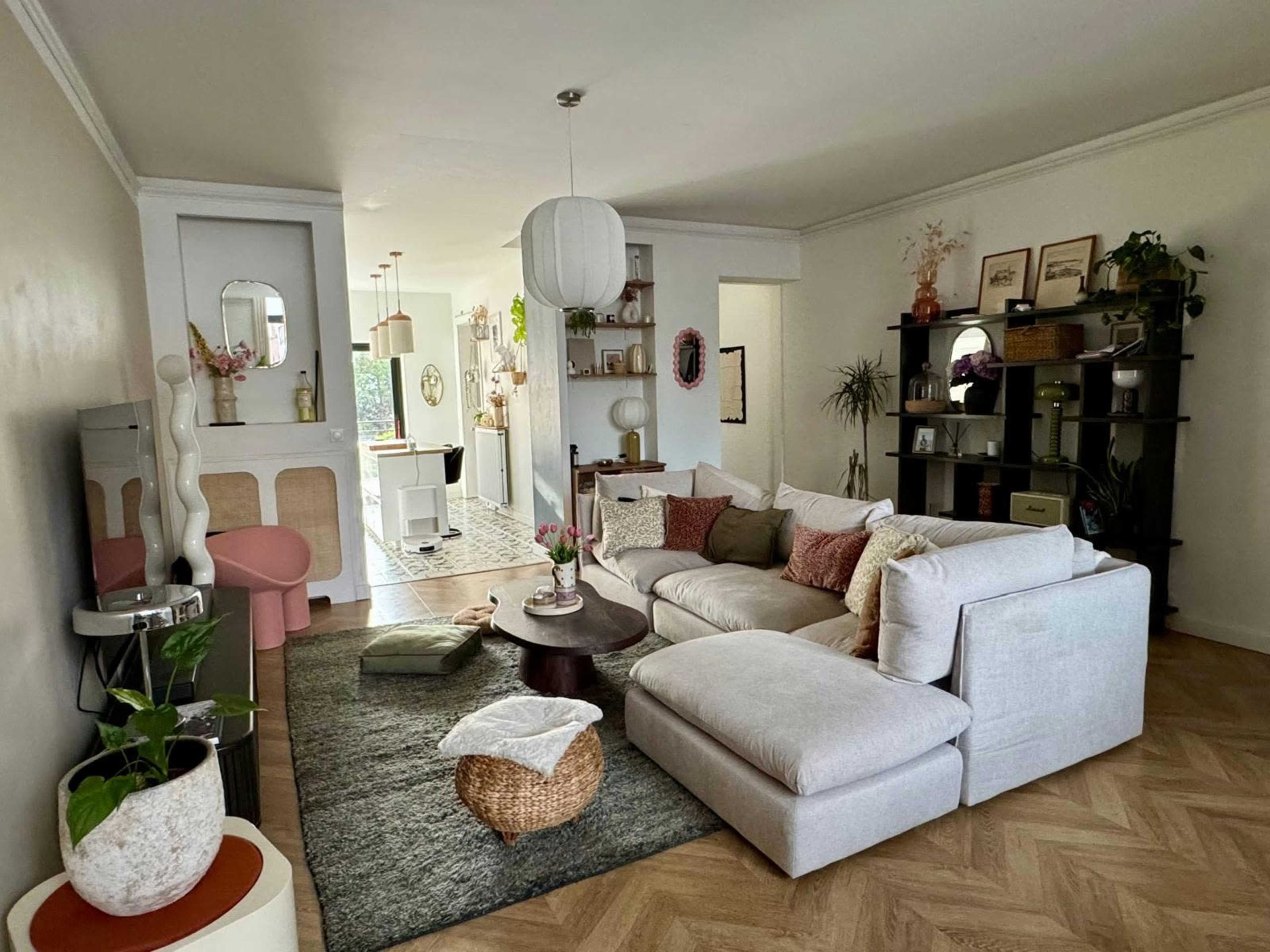 The image shows a modern living room with a large sectional sofa, a round coffee table, and decor elements such as plants and artwork against light-colored walls.