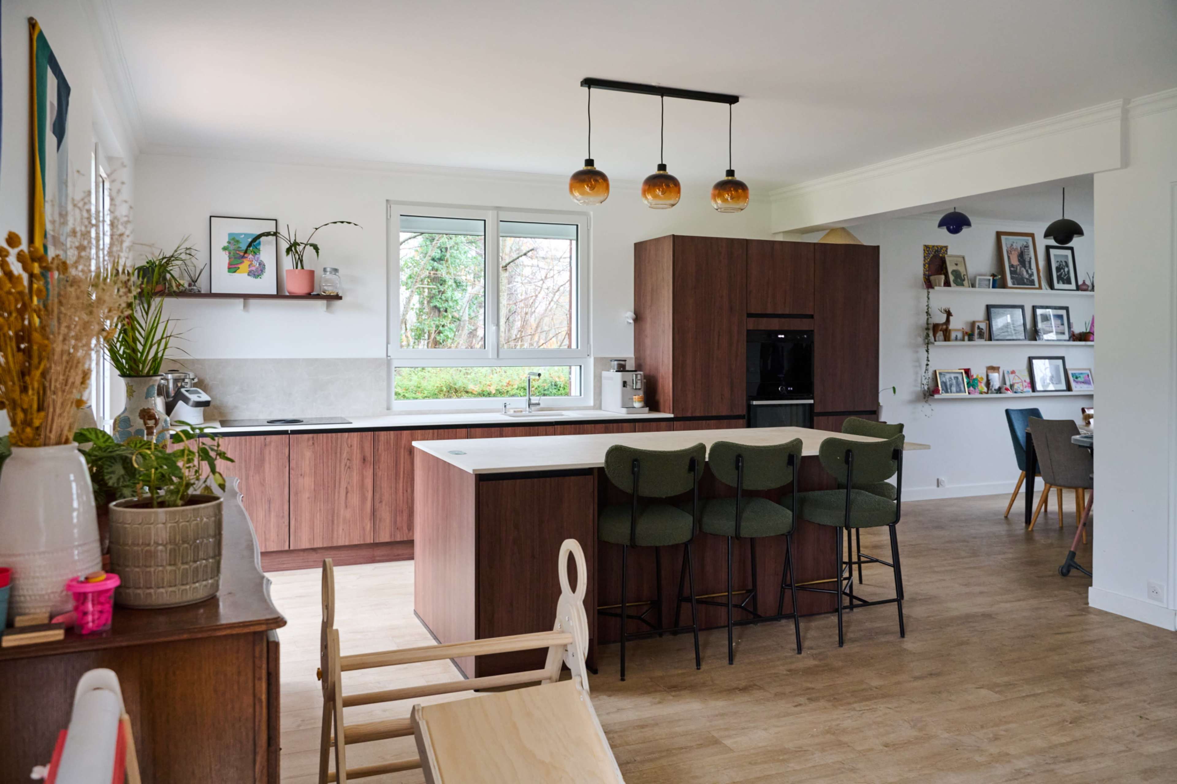 The image shows a modern kitchen with wooden cabinetry, a central island, and a dining area in an open-concept space, featuring large windows that allow natural light to illuminate the room.