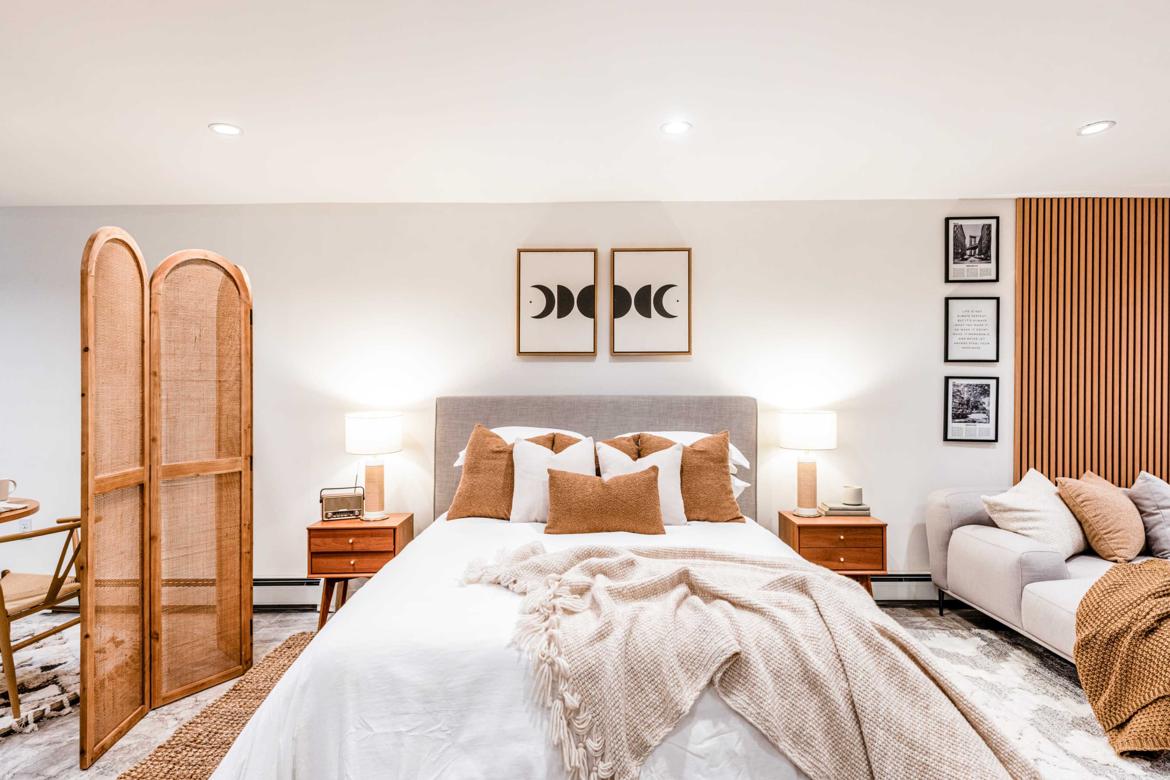 The image shows a cozy bedroom featuring a neatly made bed, a wooden room divider, two bedside tables with lamps, and a seating area with a light-colored couch.
