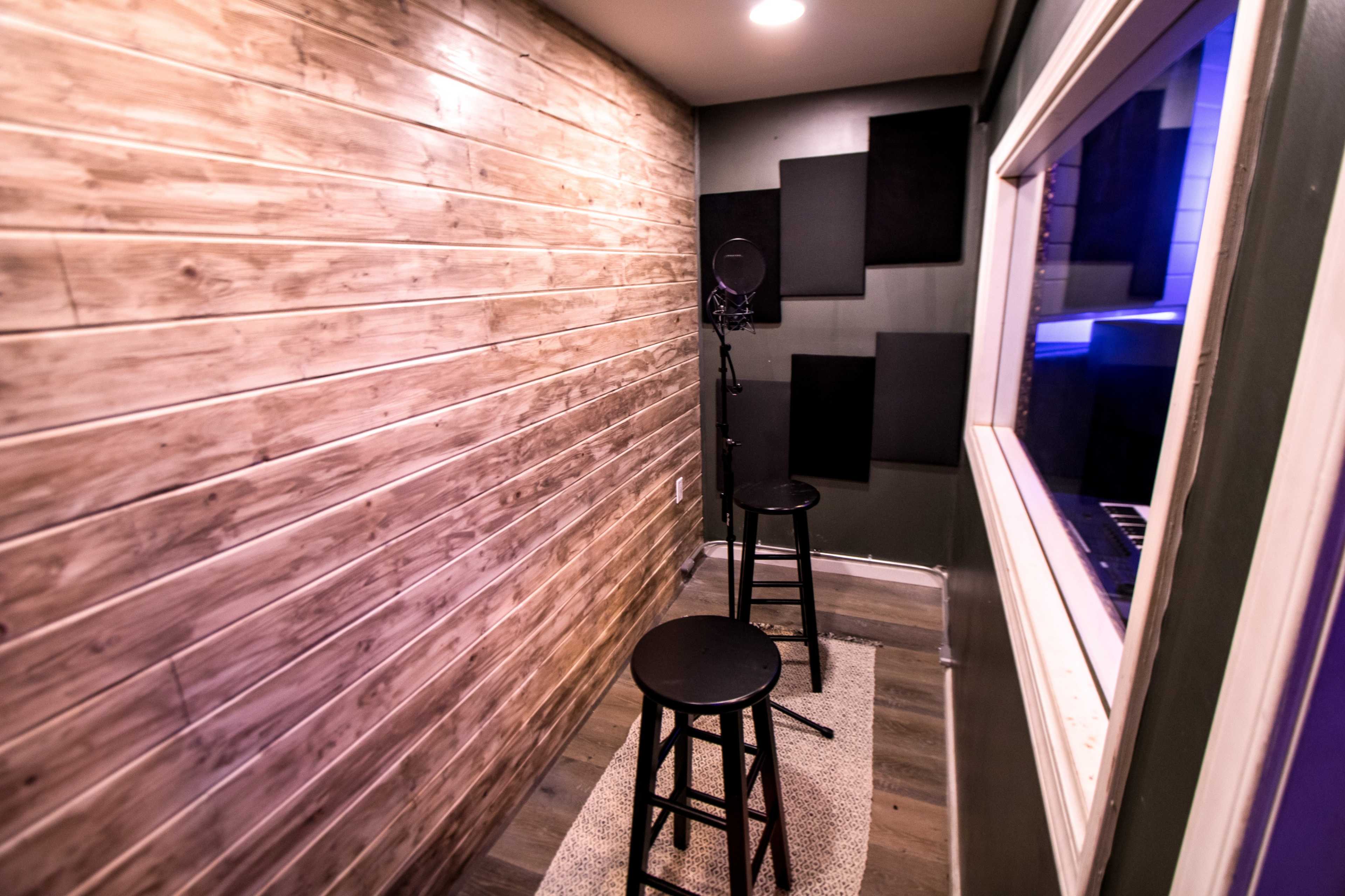A small recording studio booth with a textured wooden wall, two black stools, and a microphone stand.
