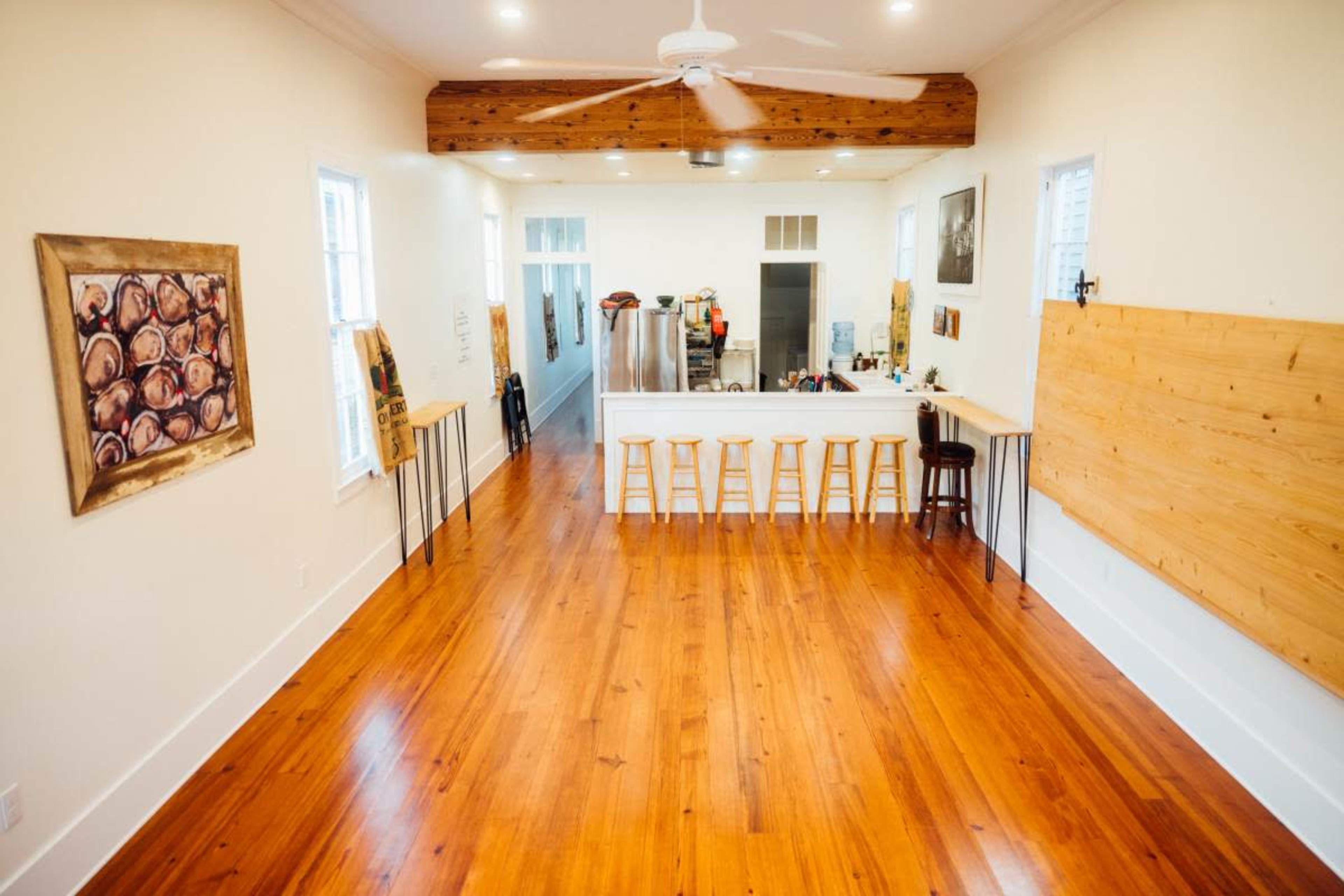 A spacious, well-lit room featuring polished wooden floors, a white kitchen island with stools, and a ceiling fan above.