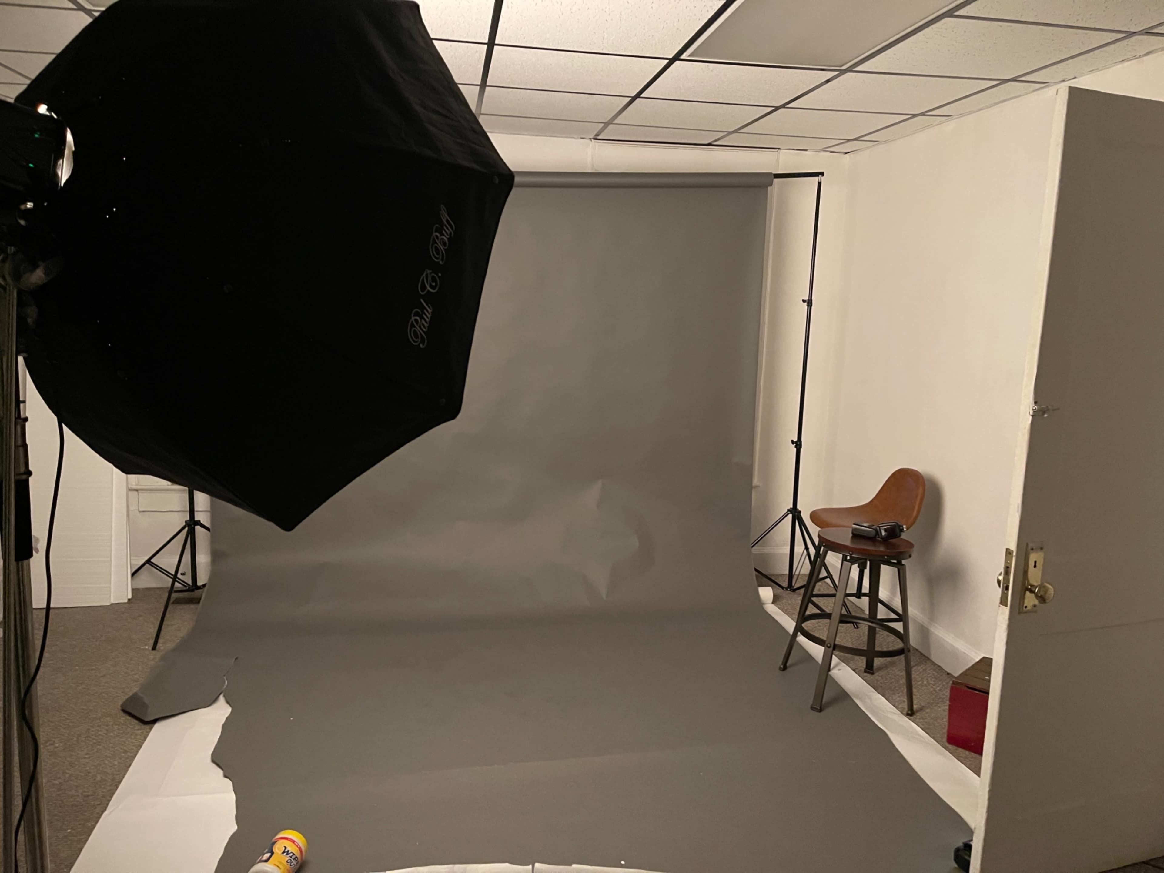 A photography studio with a gray backdrop, lighting equipment, and a stool positioned against a plain wall.