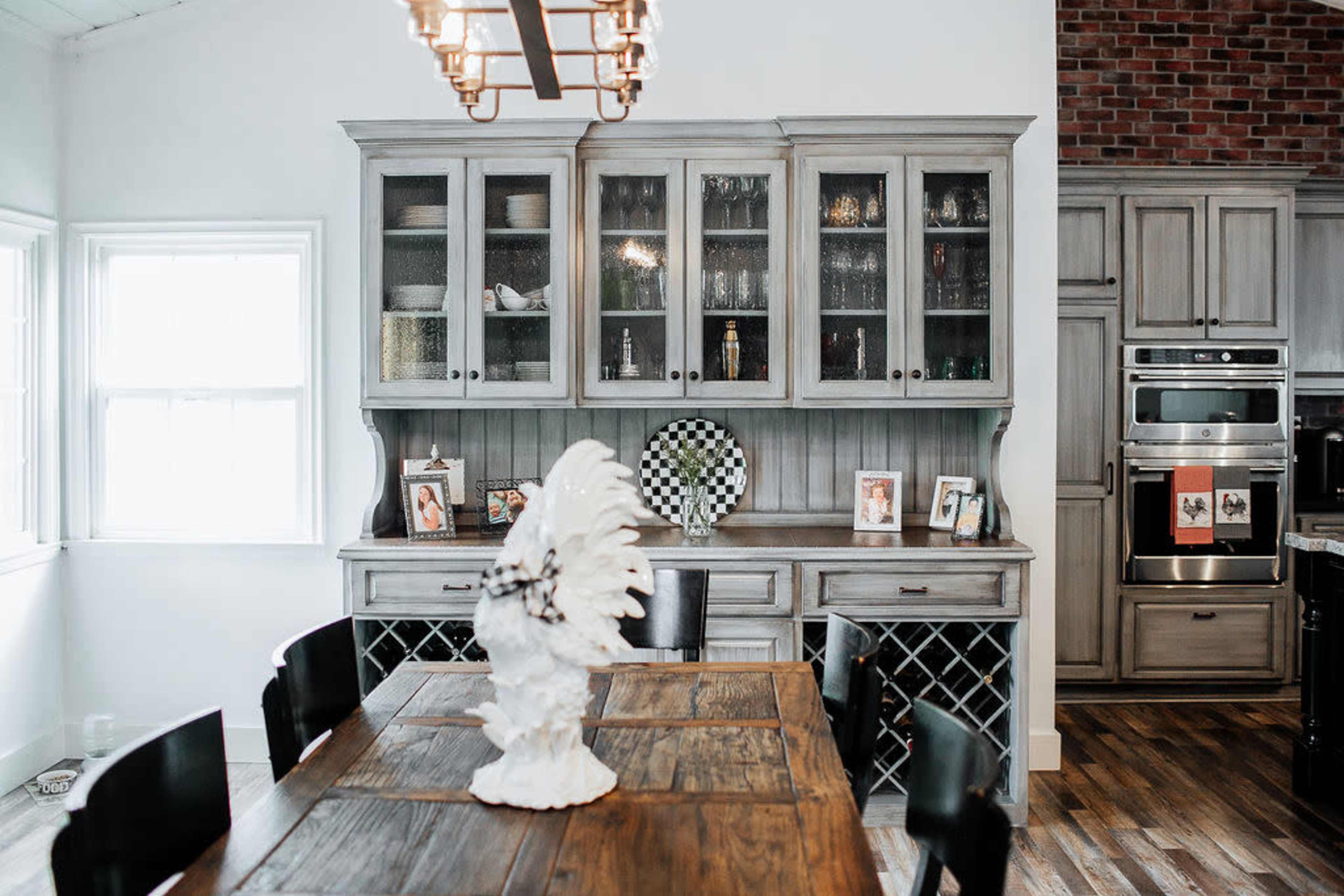 A modern kitchen featuring a wooden dining table, a hutch with glass doors displaying tableware, and a brick accent wall.
