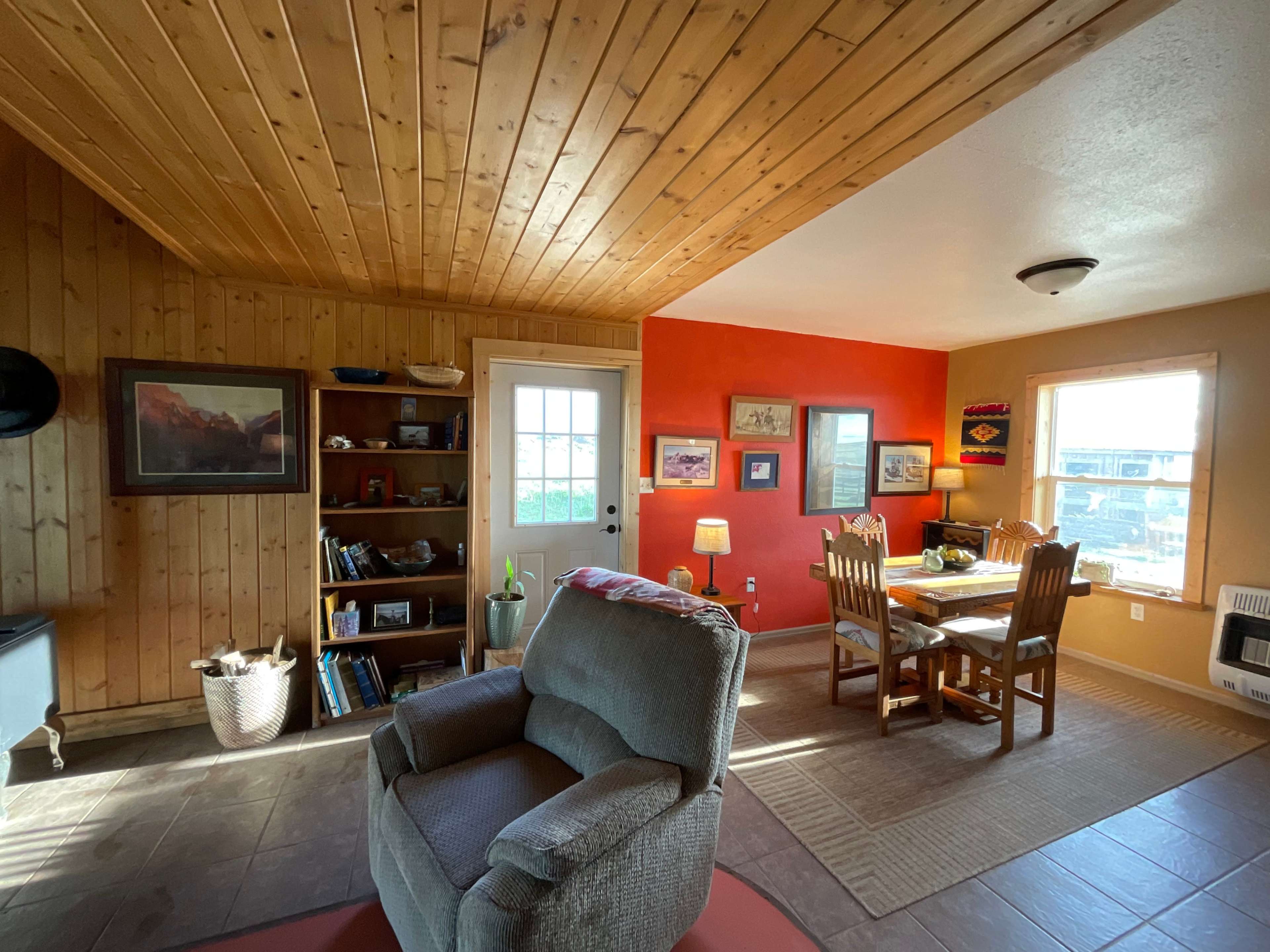 The image shows a cozy living space featuring wooden walls, a dining area with a table and chairs, and a comfortable armchair positioned near a door that leads outside.