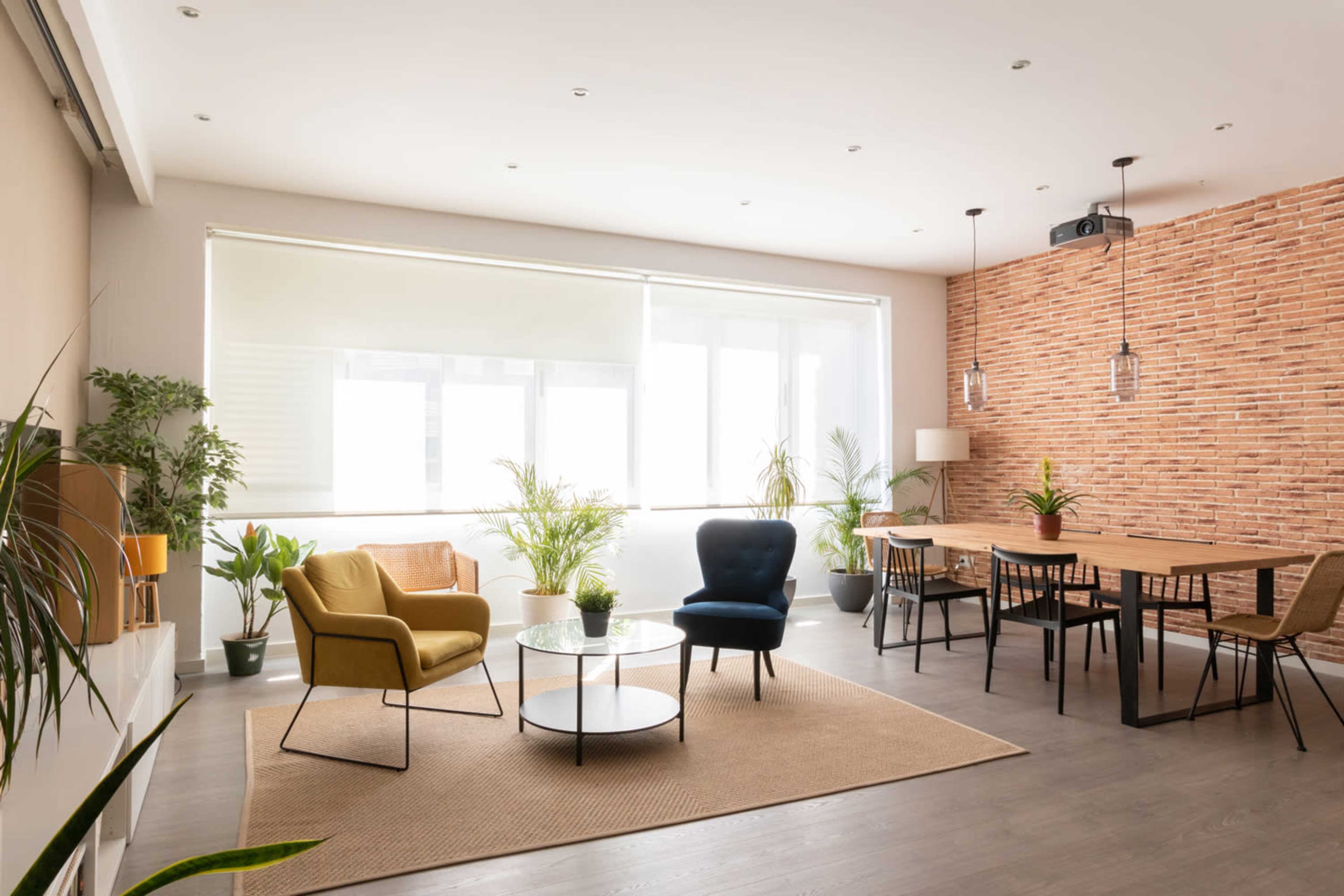 The image shows a modern living space featuring a beige and brick wall, a round table with chairs, and various plants arranged around the room.