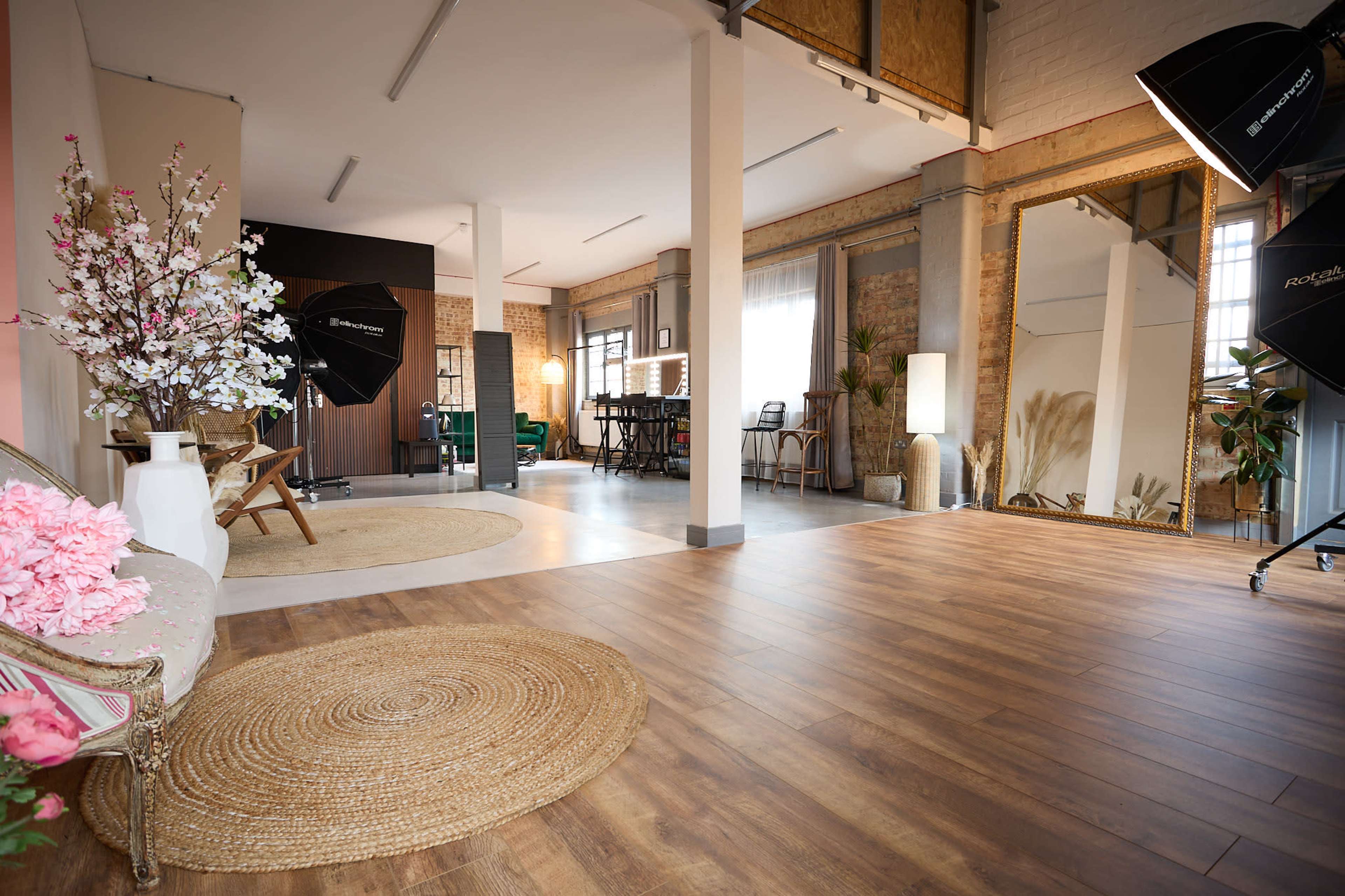 The image shows a spacious loft-style studio with wooden flooring, large windows, and various seating areas, including a vintage couch and a dining table.