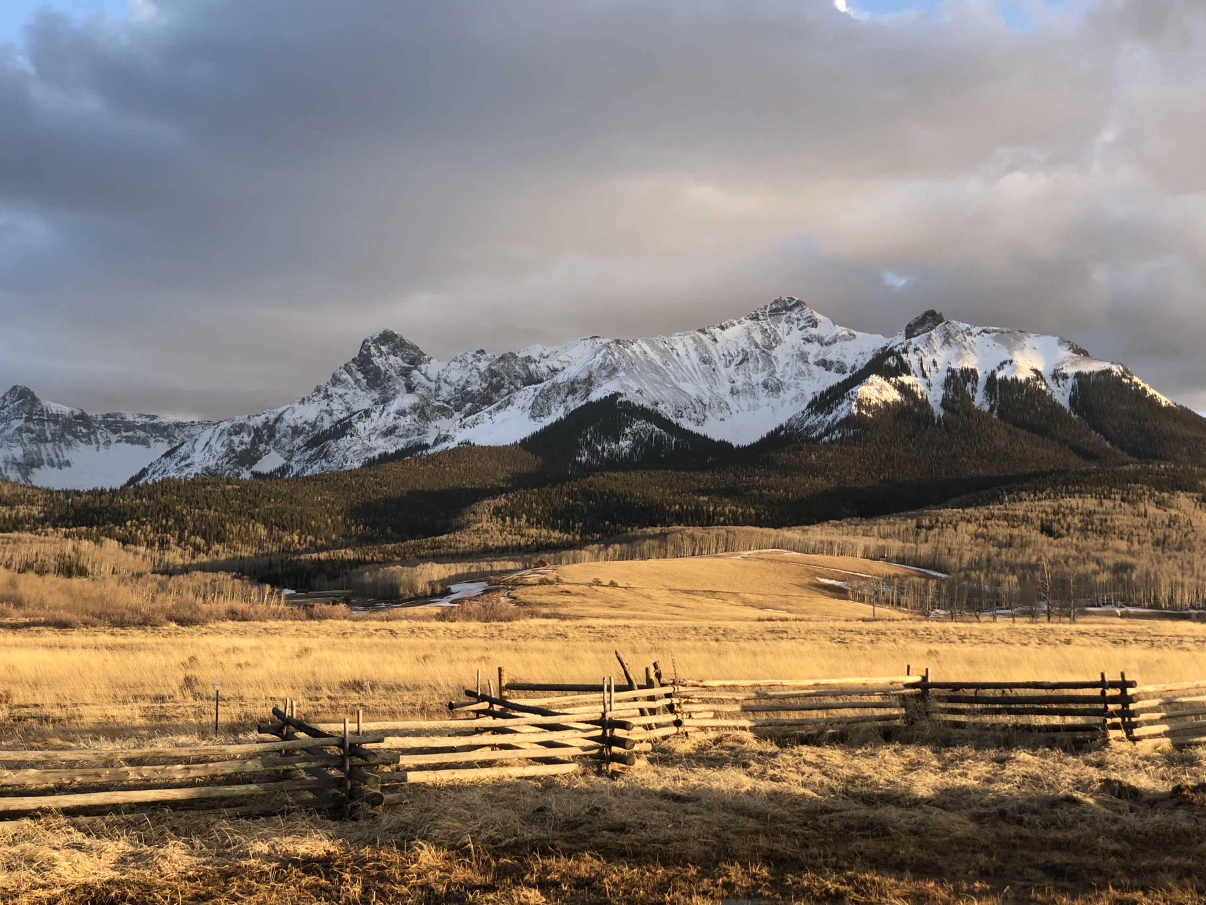 The image shows a mountainous landscape with snow-capped peaks rising above a grassy field, framed by a wooden fence in the foreground.