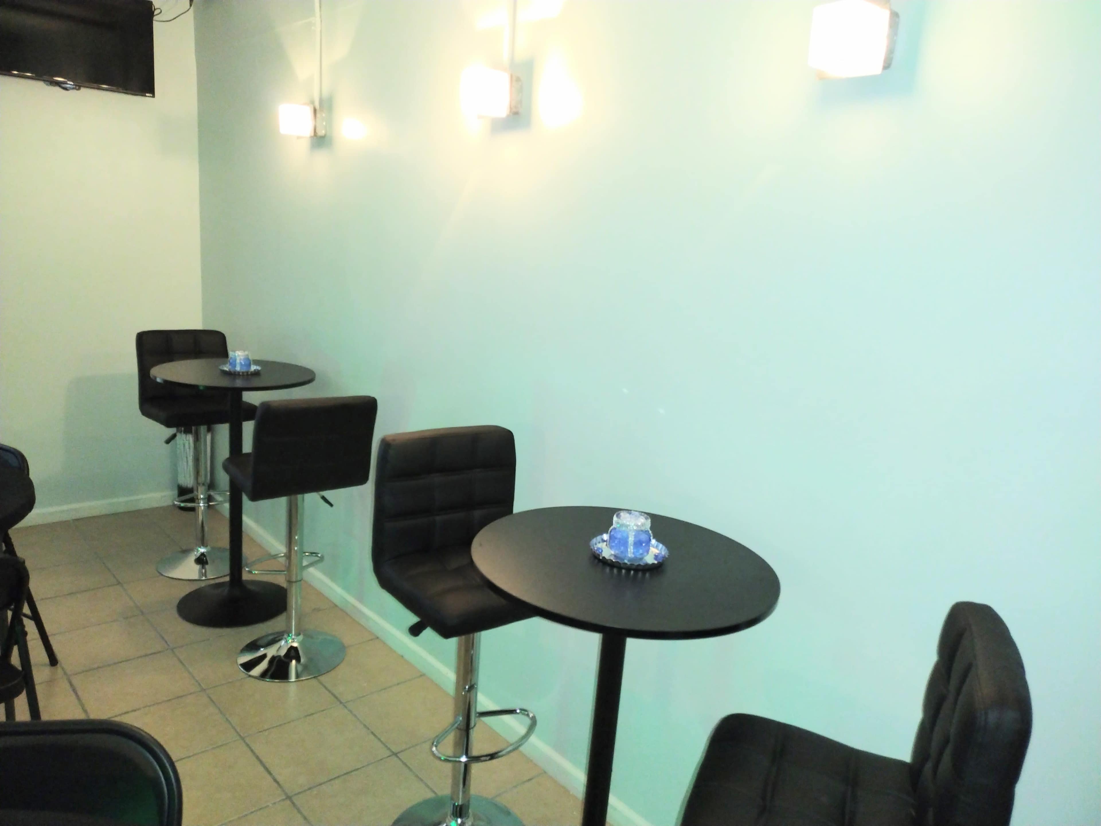 The image shows a minimalist indoor space with two black bar tables and stools against a light blue wall, illuminated by wall-mounted lights.