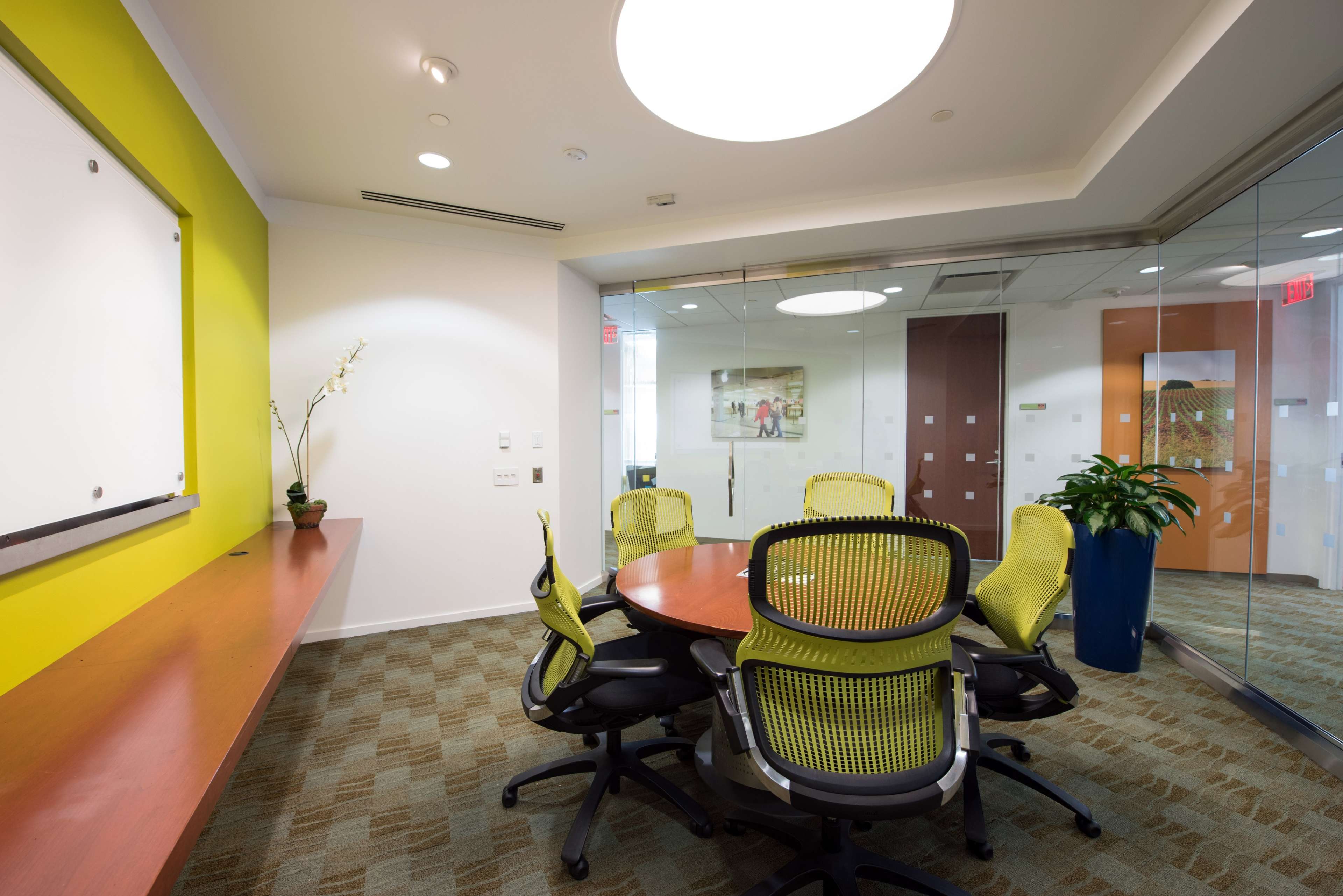 The image shows a modern conference room featuring a round table surrounded by ergonomic chairs, with a whiteboard on one wall and glass partitions.