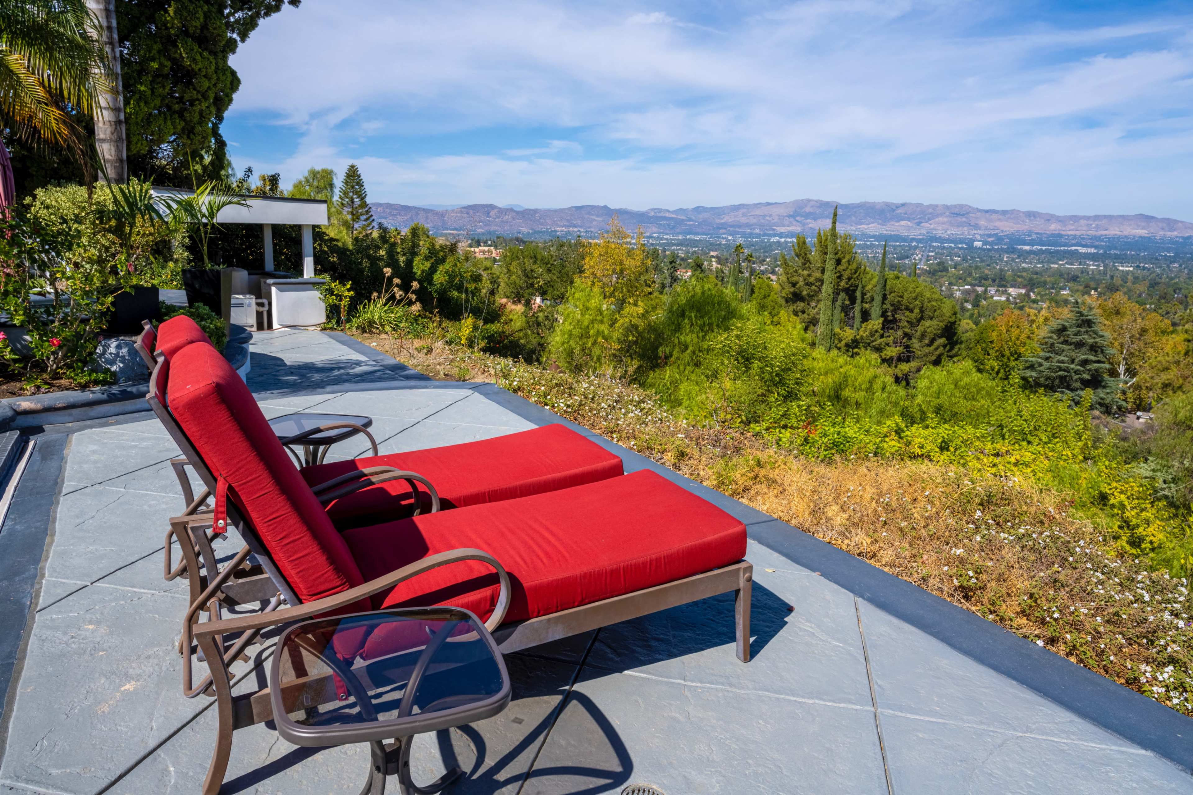 Two red lounge chairs are positioned on a stone patio overlooking a valley filled with greenery and distant mountains under a blue sky.