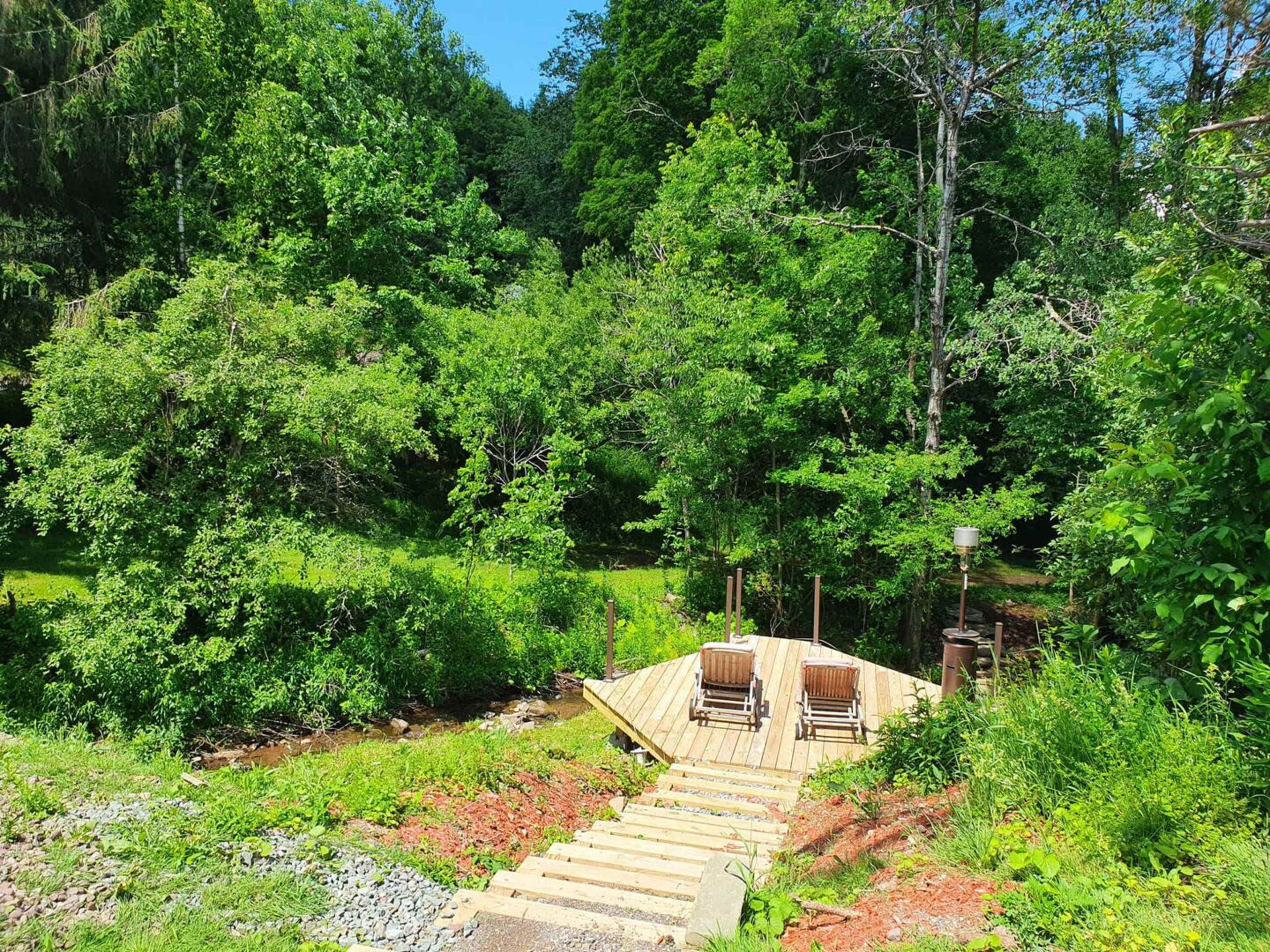 A wooden deck with two chairs overlooks a lush green forest and stream.