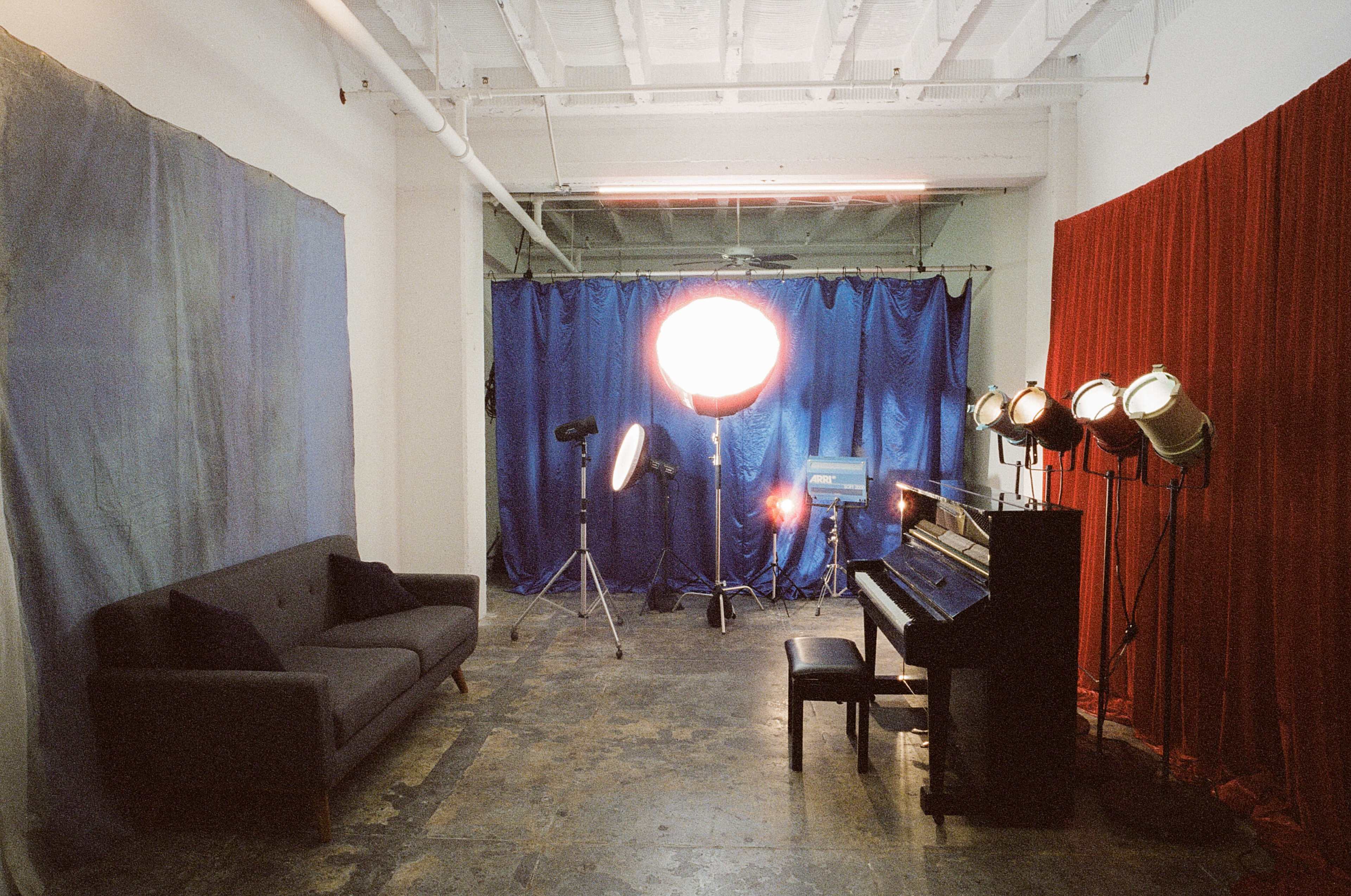 A studio space with a gray sofa, a piano, and various lighting equipment set against blue and red curtain backdrops.
