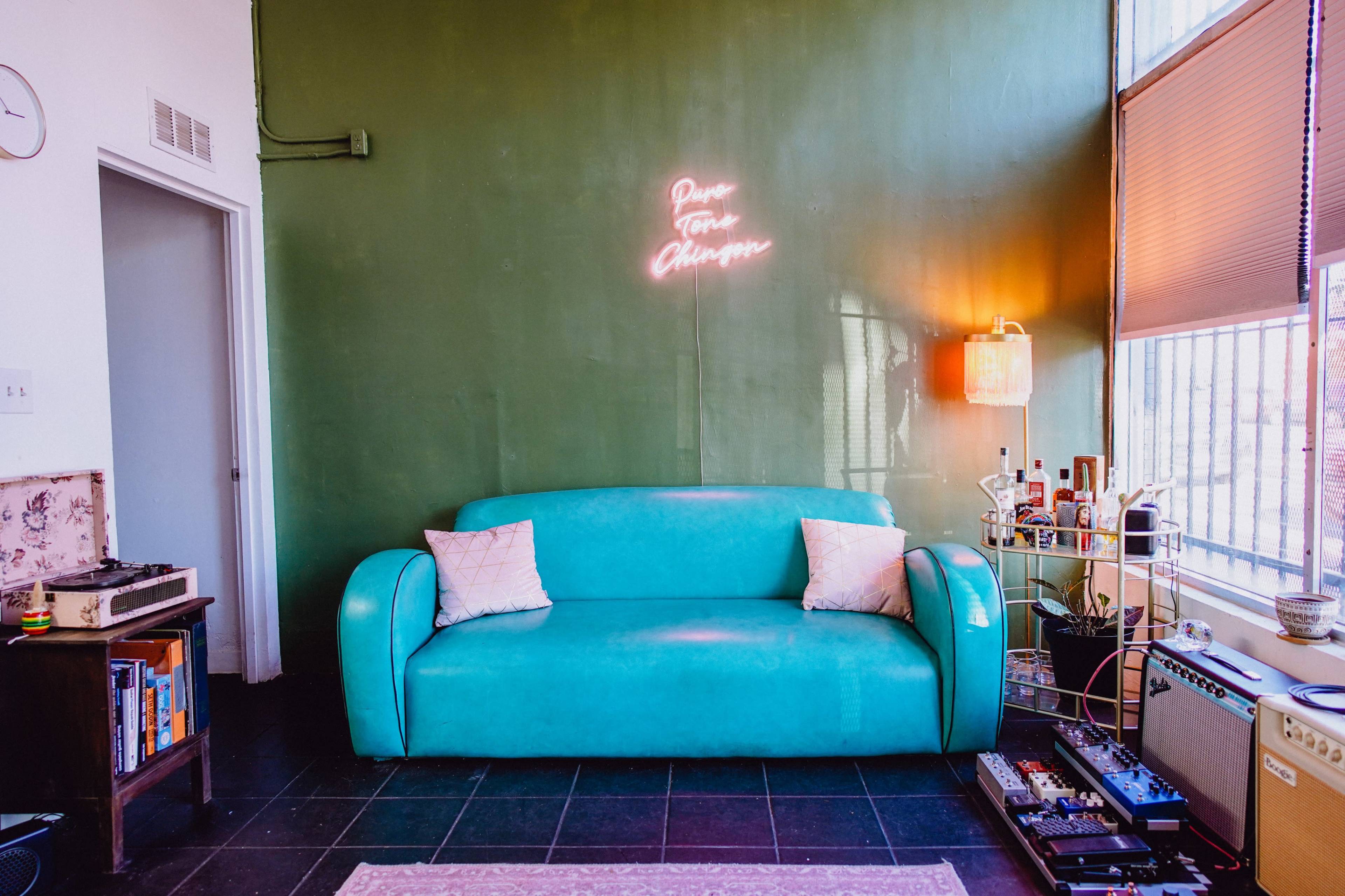 A teal sofa is positioned against a green wall with a neon sign, while nearby furniture and decor create a cozy living space.