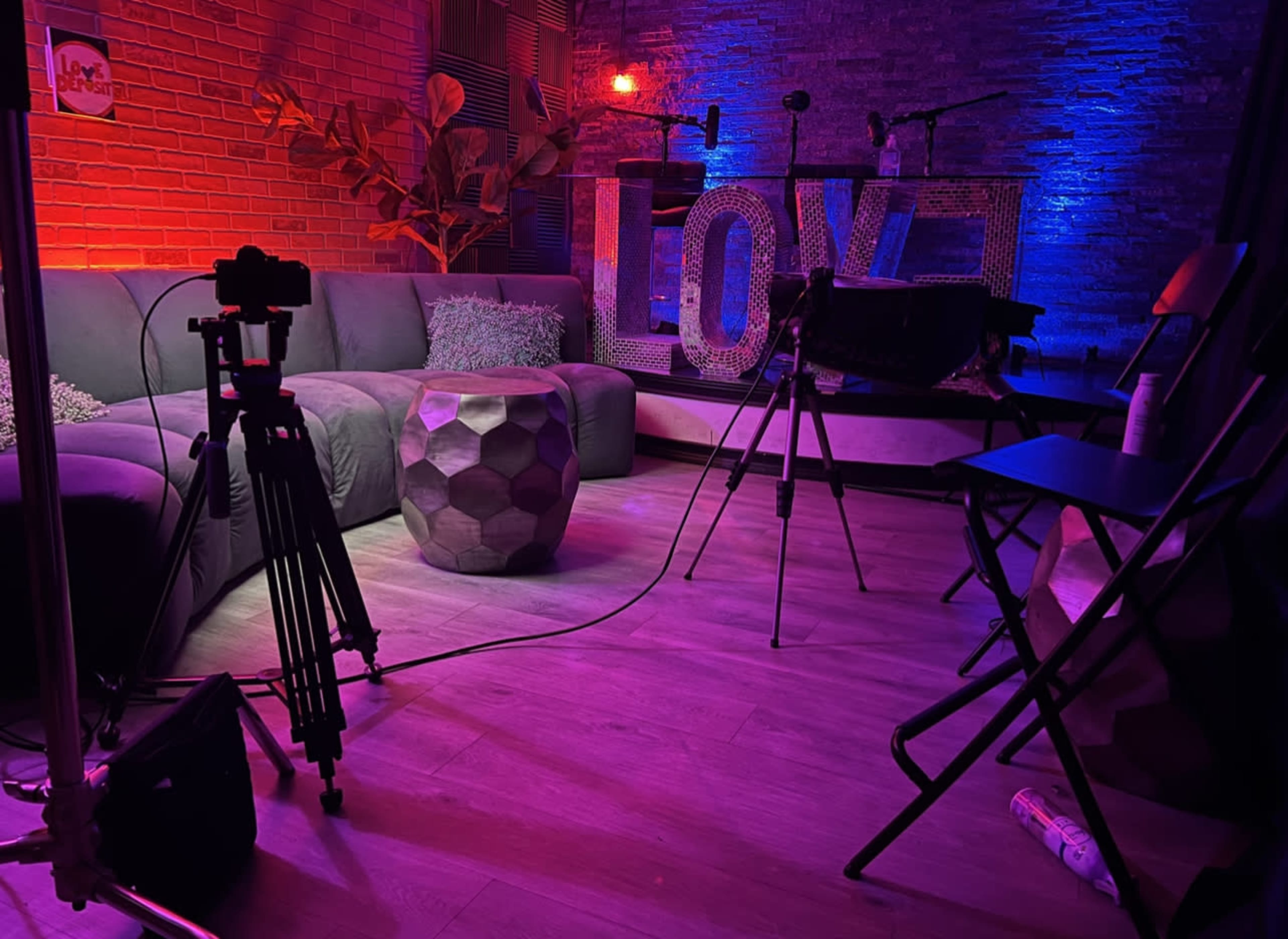 The image shows a dimly lit studio setup with a green sofa, a geometric coffee table, and a large "LOVE" sign illuminated in the background.