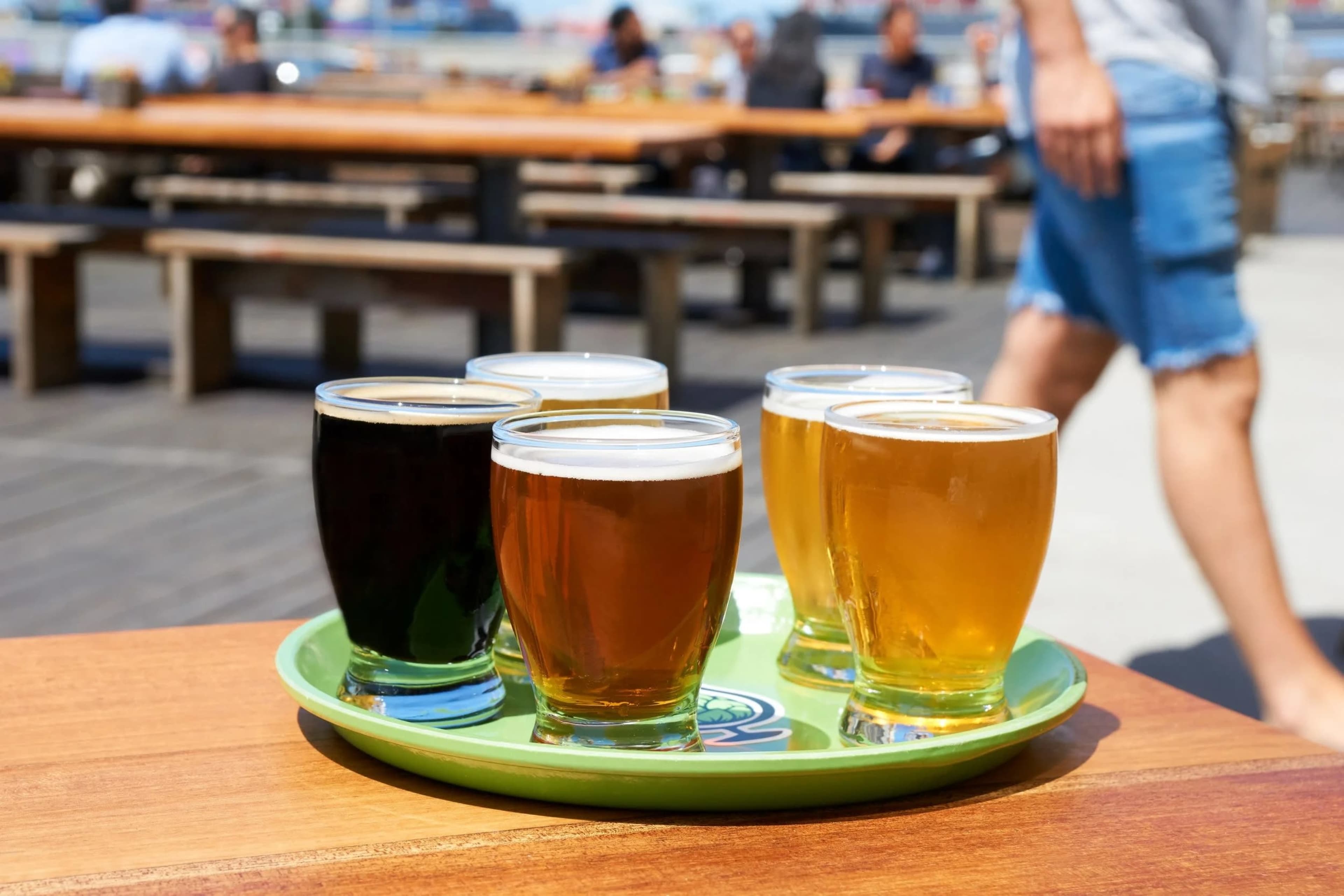 A tray holds six glasses of beer in various colors, placed on a wooden table with a blurred background of people and seating areas.