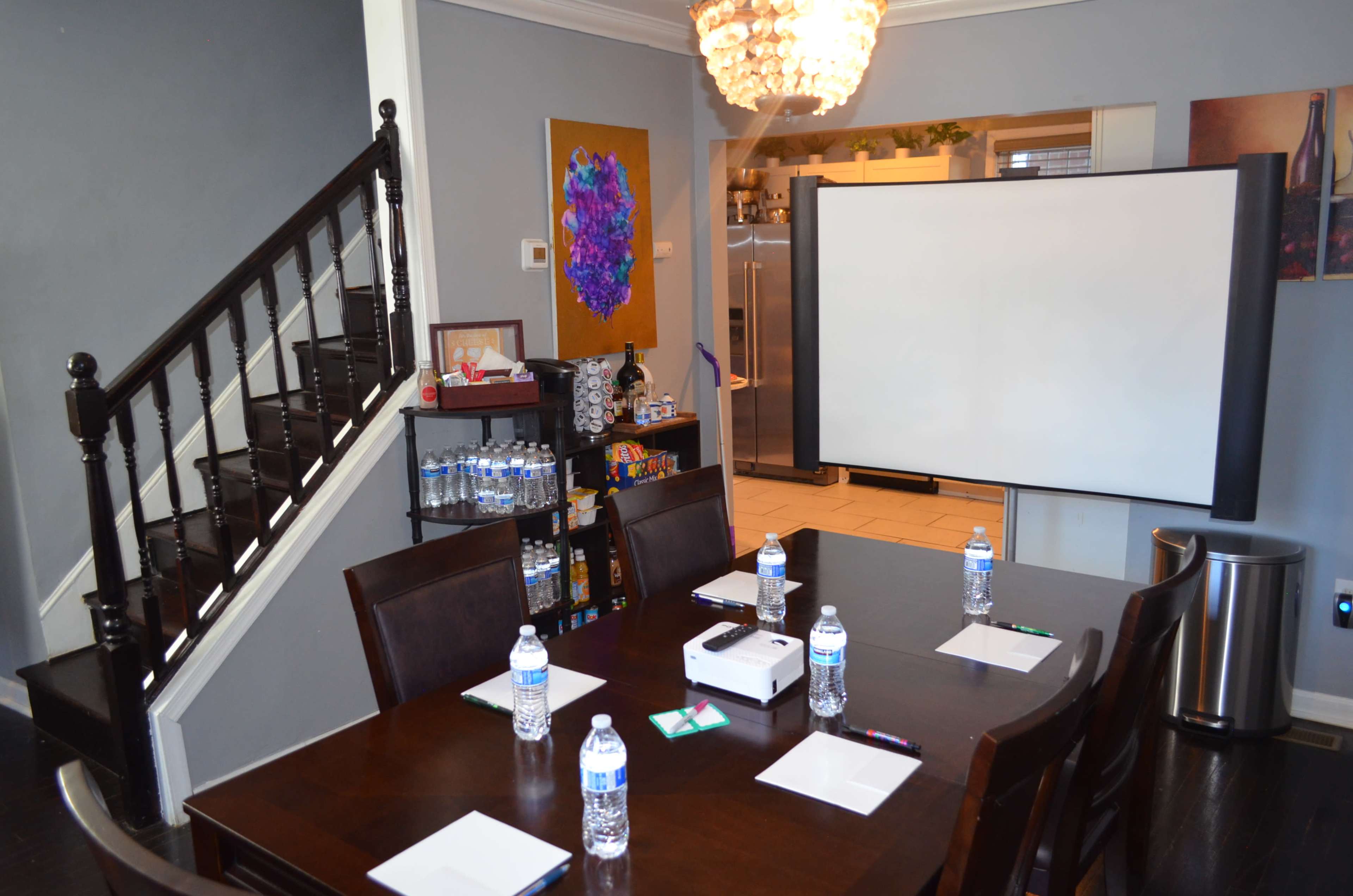 A meeting room features a large table set with water bottles, notepads, and pens, while a projection screen stands against the wall and stairs lead to an upper level.