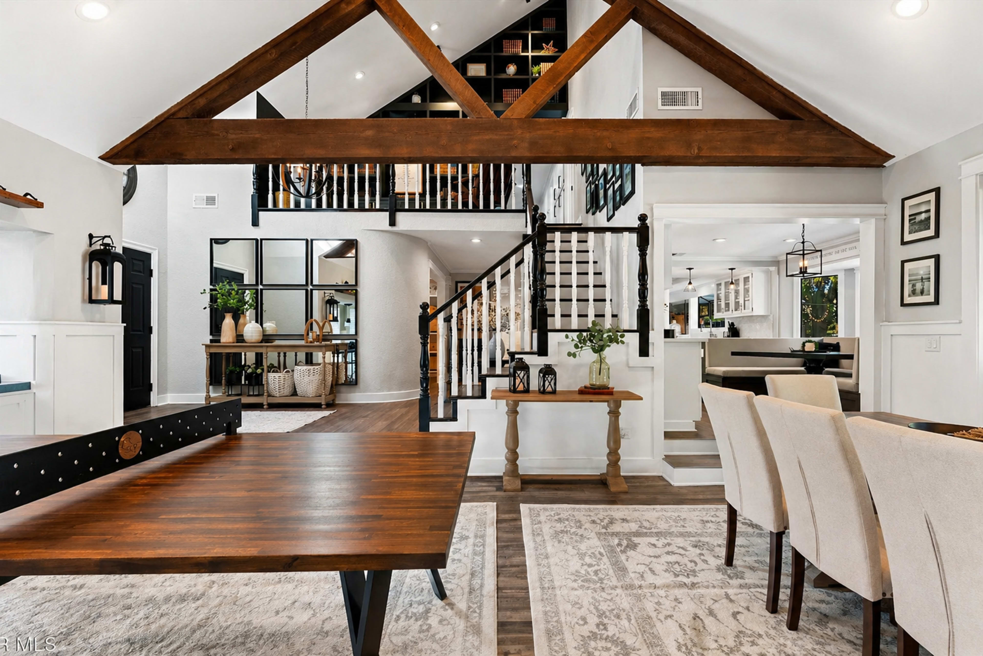 The image depicts a spacious, modern living area featuring a wooden dining table, a ping pong table, and a staircase leading to an upper level with a balcony and open-concept kitchen.