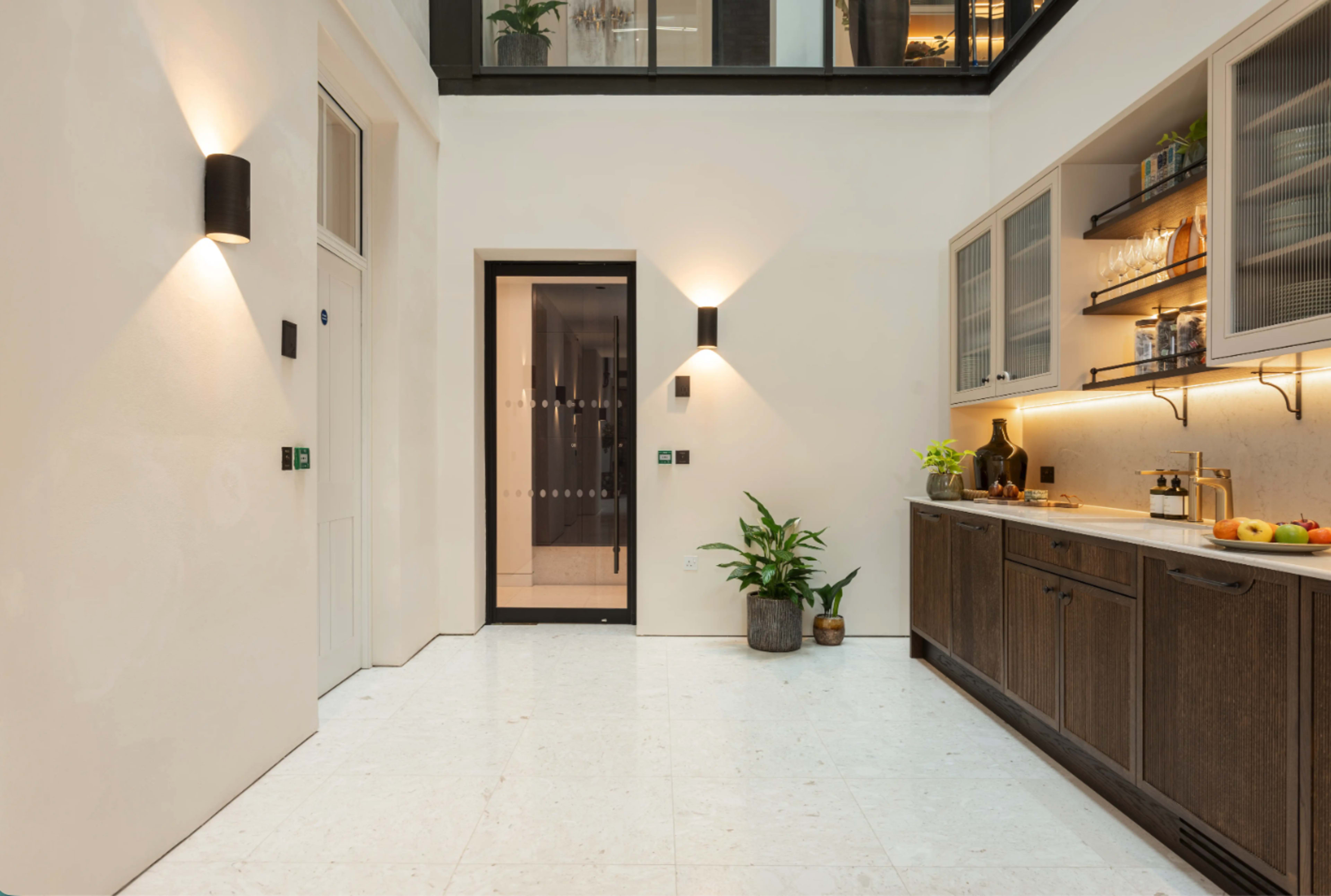 The image shows a modern kitchen area with wooden cabinetry, a door leading to another space, and a potted plant against a light-colored wall.
