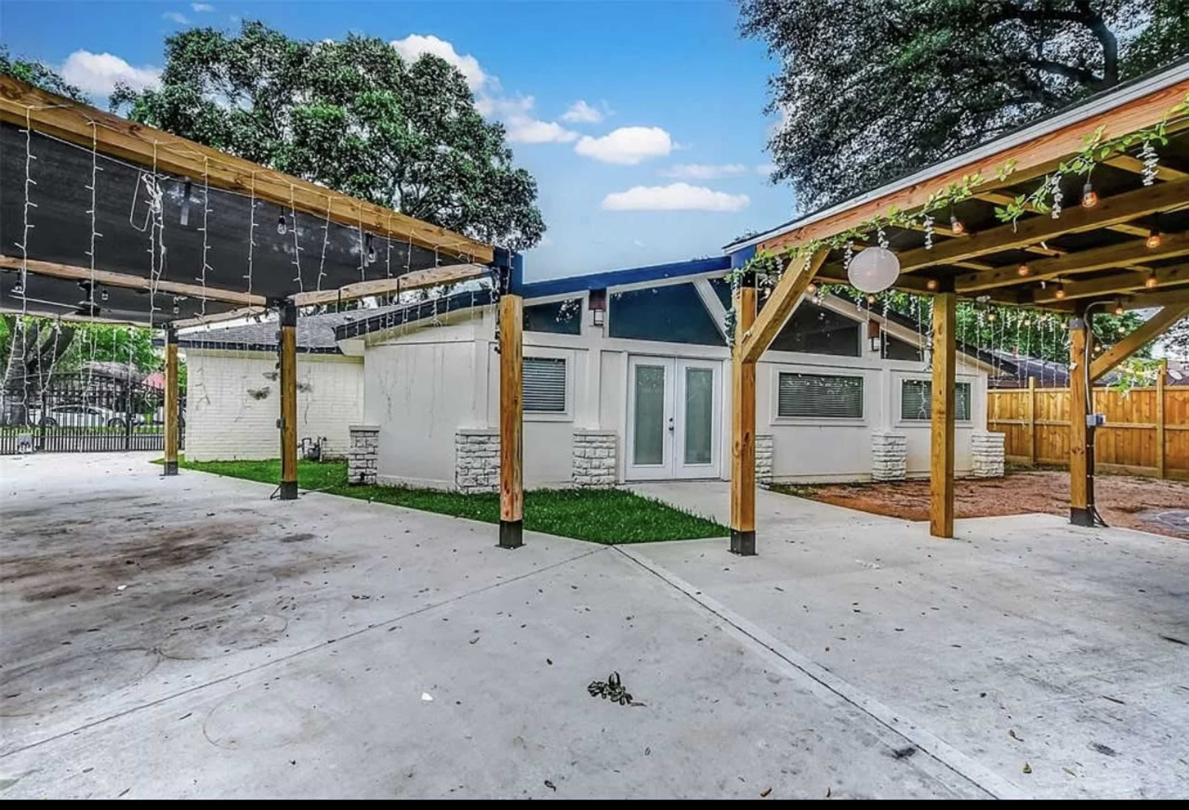 The image shows a modern house with a white exterior, surrounded by wooden pergolas and a concrete patio.