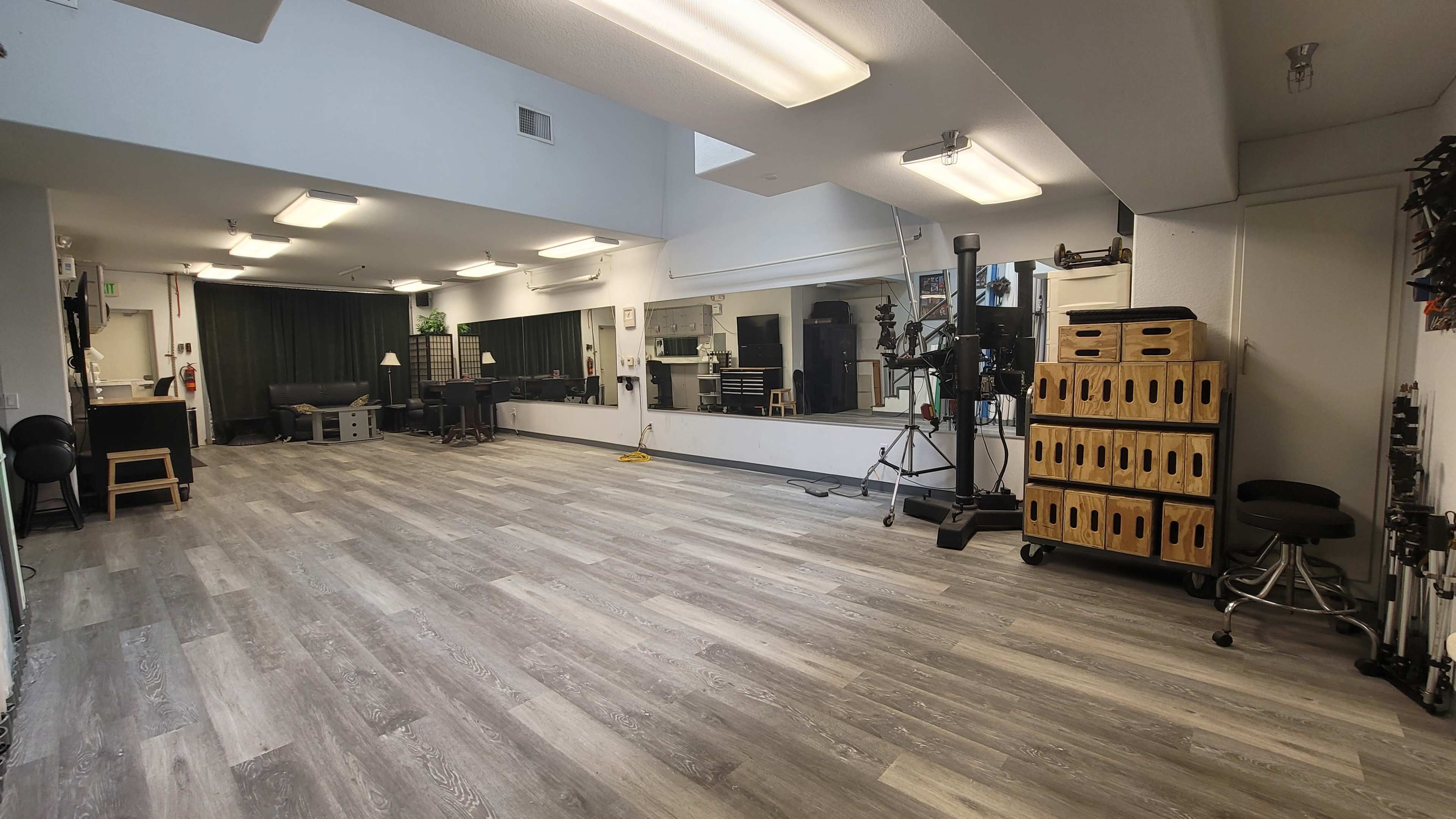 A large, empty room with gray wood flooring, equipped with mirrors, studio lighting, and storage boxes along the wall.