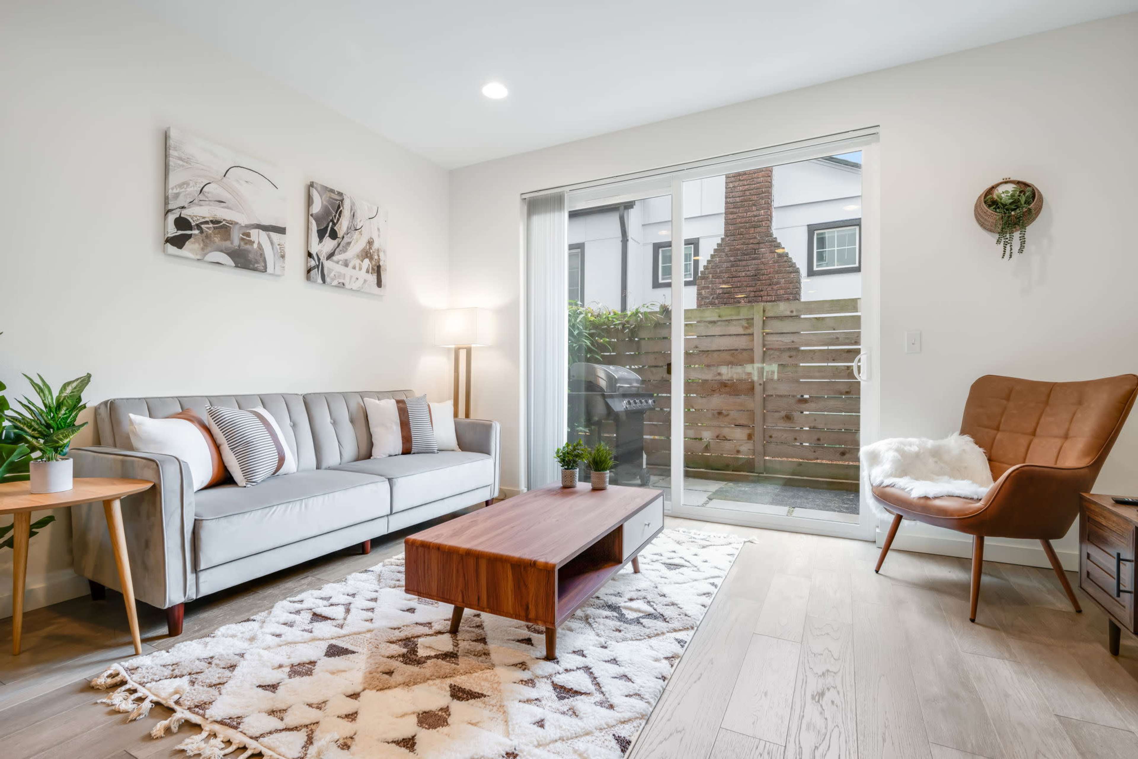 A modern living room features a light gray sofa, a wooden coffee table, and a plant, with a glass door leading to a small outdoor area.