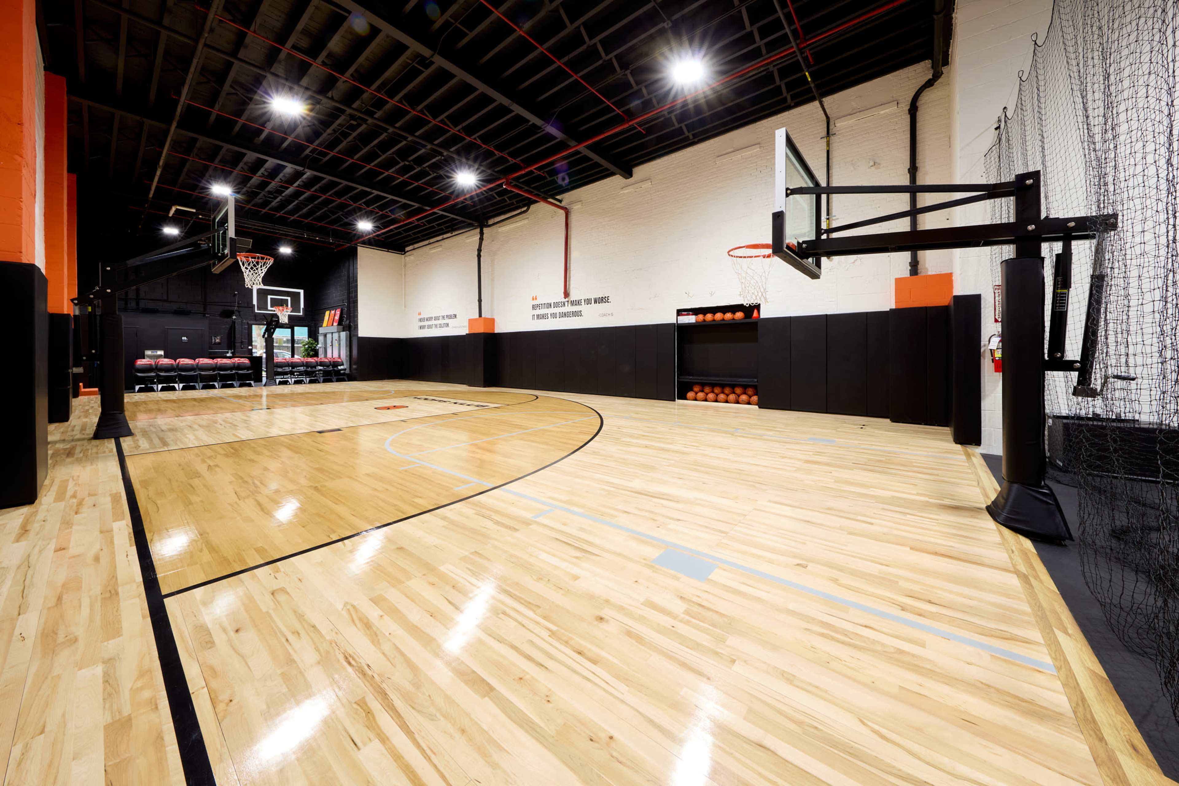 The image shows a well-lit indoor basketball court featuring polished wooden flooring, basketball hoops, and seating along one side.