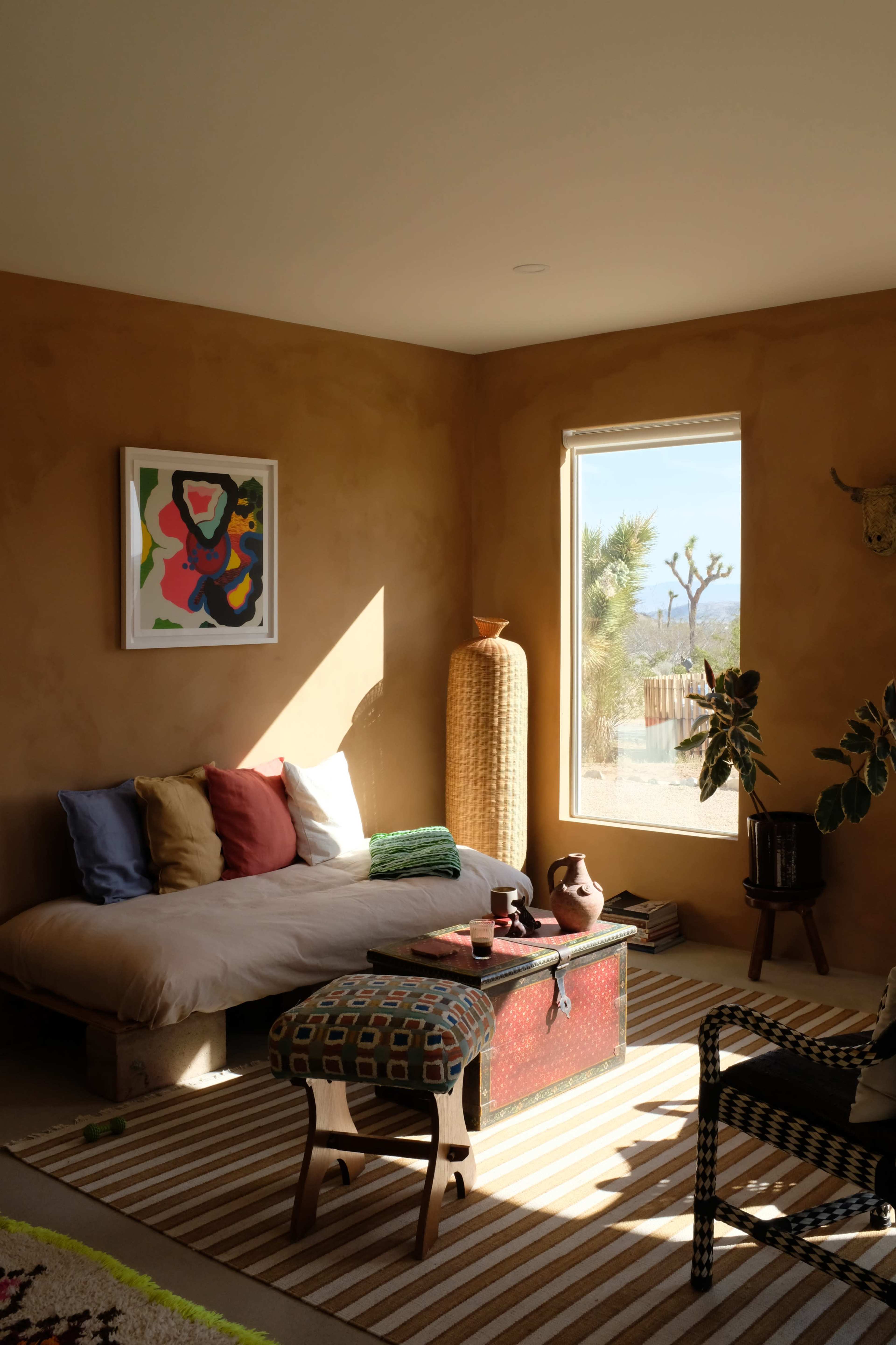 A minimalist living room features a low seating area with colorful cushions, a patterned rug, and a large window showcasing a desert view.